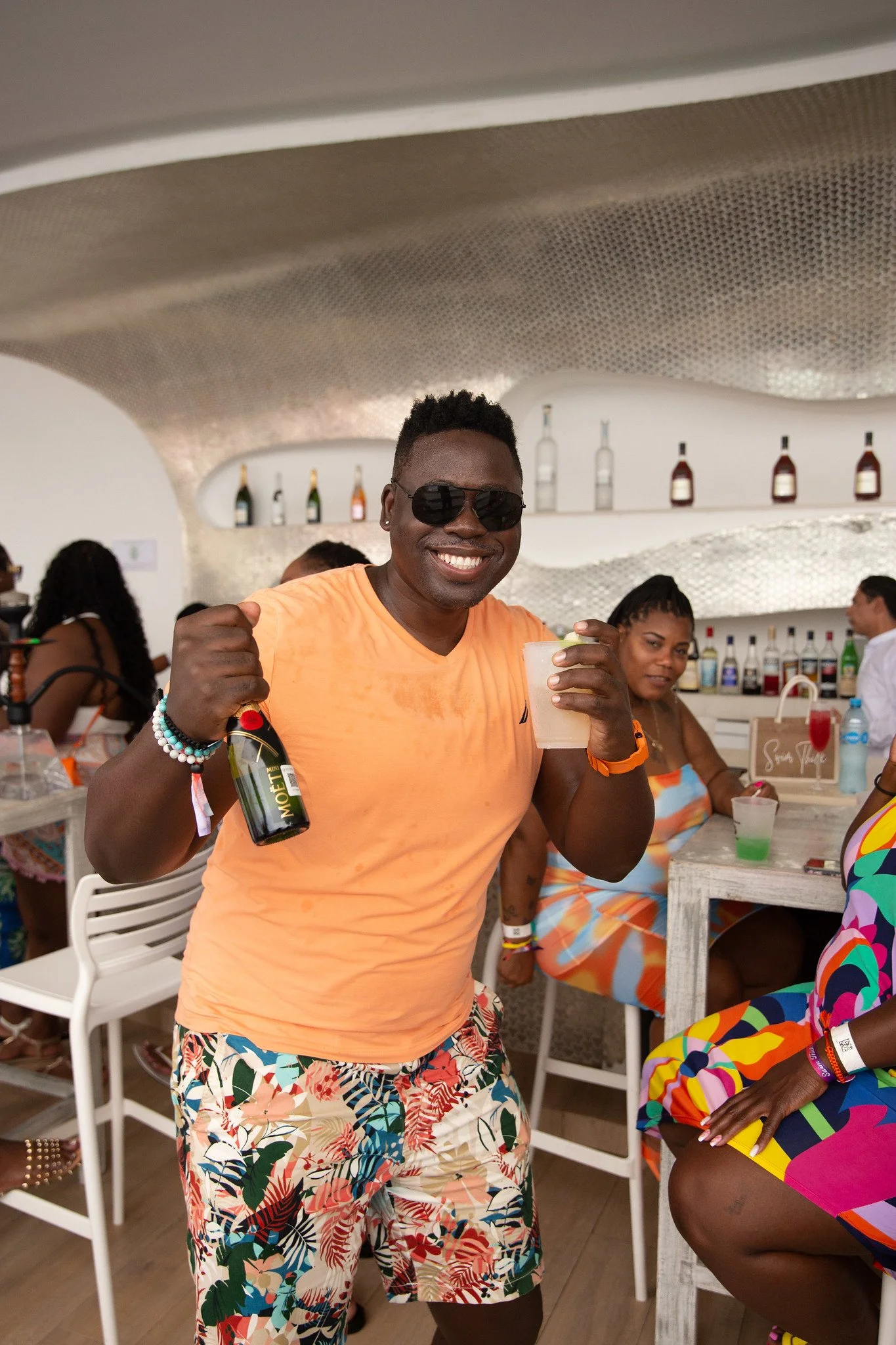 Happy man wearing sunglasses and tropical shorts holding a drink and a bottle at a lively poolside or beach bar party.