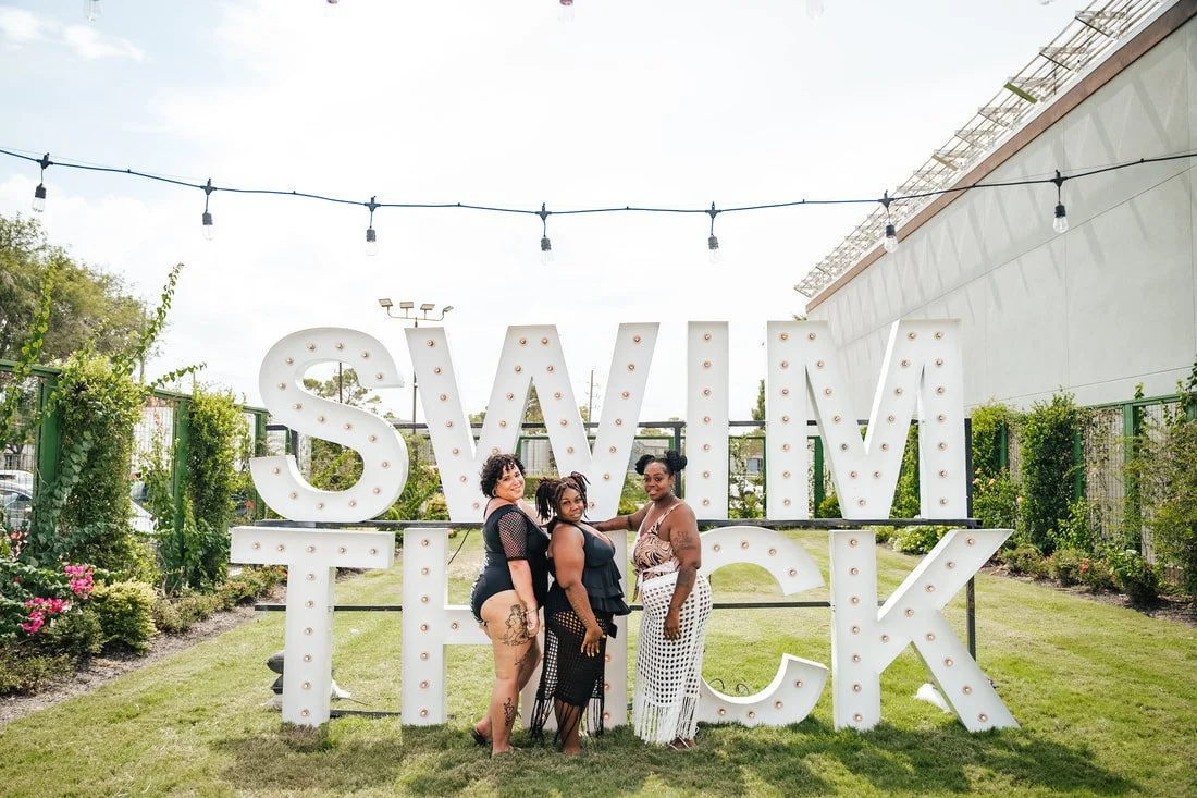Three women standing in front of large illuminated letters spelling "SWIM, FIT, LICK" in an outdoor garden setting.