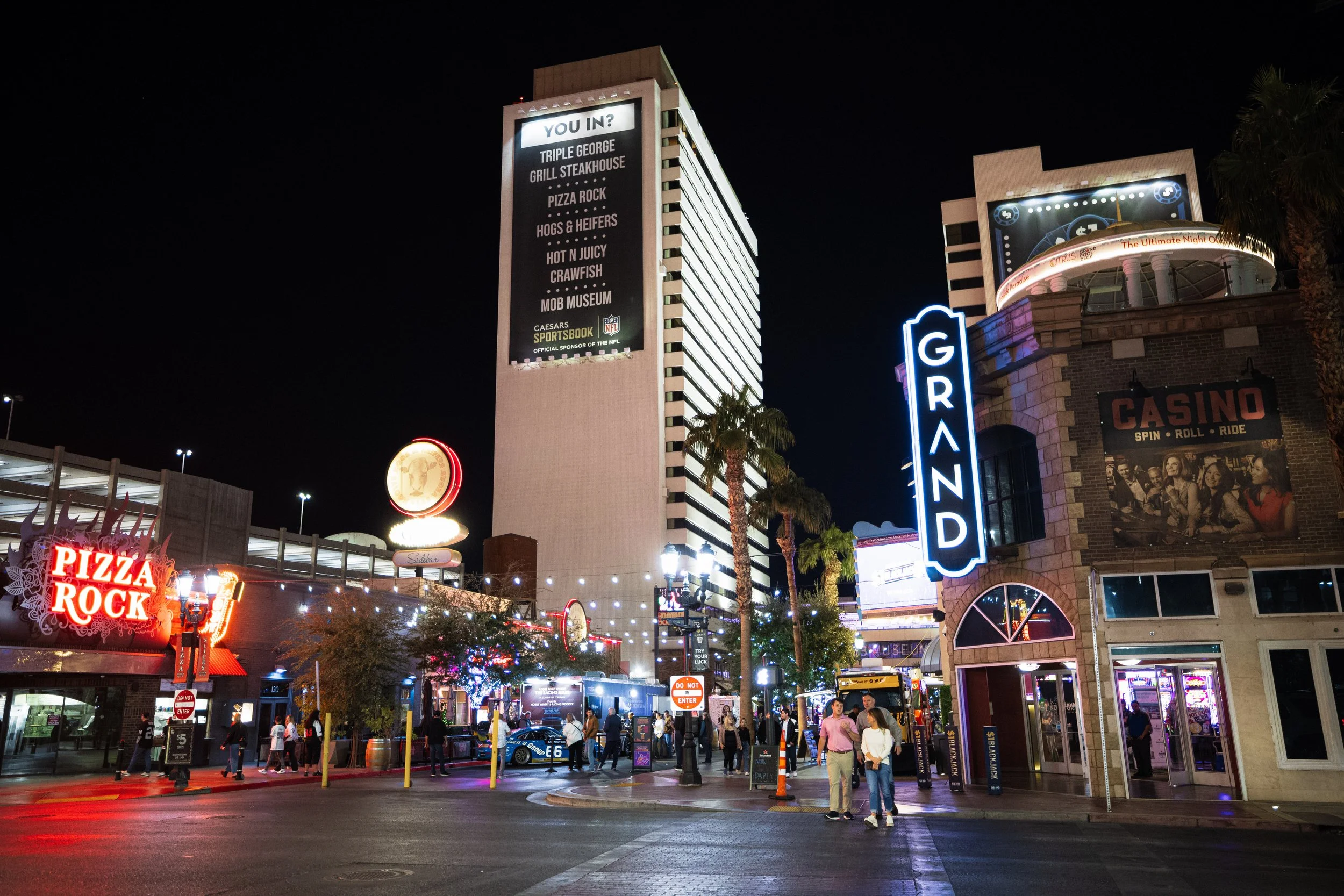 Night scene of the Las Vegas Strip with neon signs, a tall building with an electronic board, palm trees, and people walking on the street.