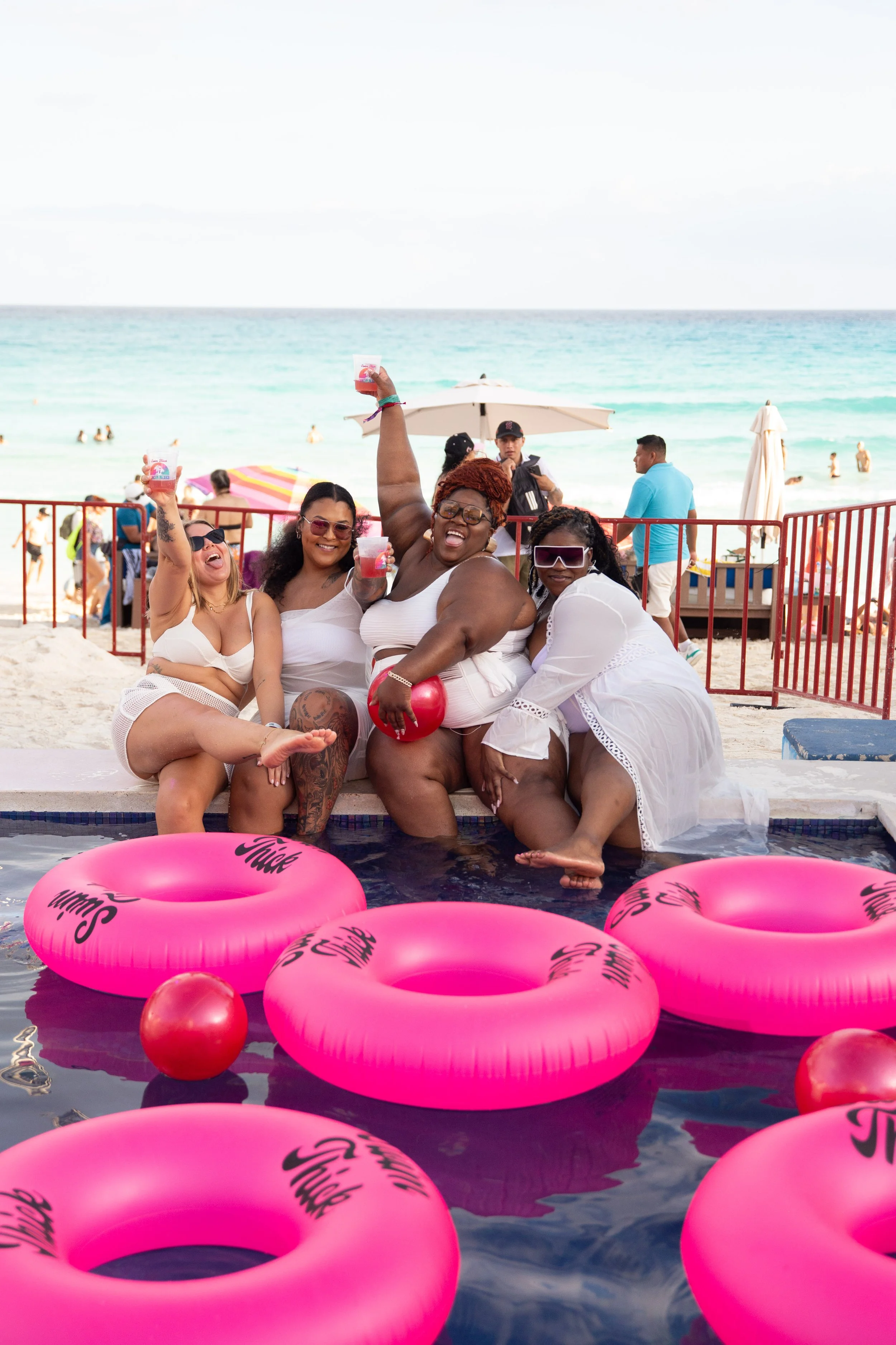 Four women enjoying a pool party at the beach, sitting on the edge of a pool with pink inflatables and holding drinks, with the ocean and beachgoers in the background.