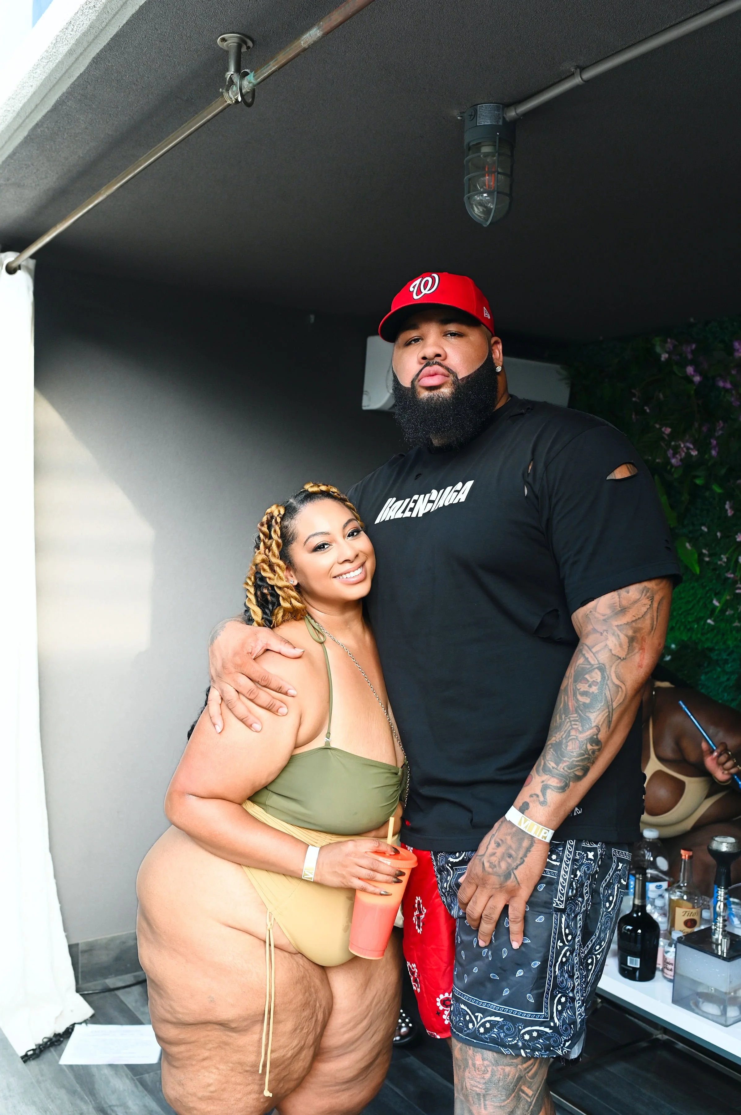 A woman in a green bikini top and yellow shorts smiling and holding a pink drink, standing next to a tall man with a beard, tattoos, wearing a black Balenciaga T-shirt and patterned shorts, with an arm around her shoulder, at an indoor party.