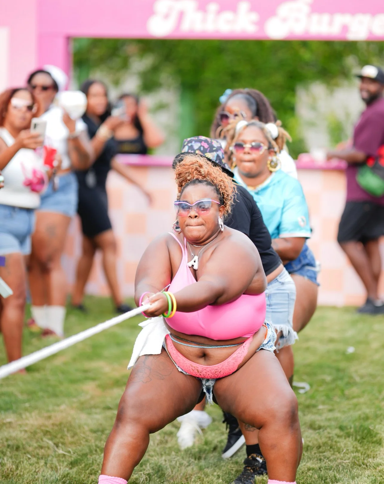 A woman in pink tank top and frayed shorts participating in a tug-of-war game at an outdoor event, with spectators watching and taking pictures in the background.