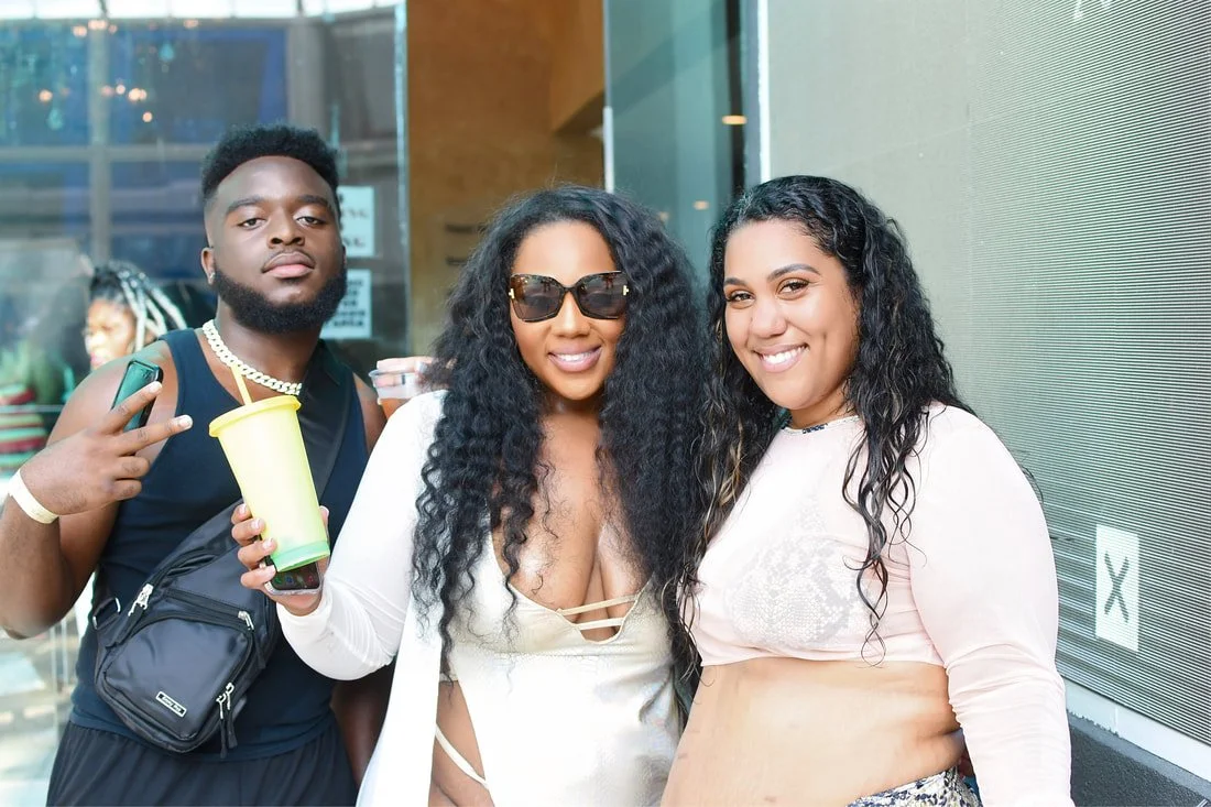 Three people smiling and posing for a photo outside a building. One man holding a drink and making a peace sign, two women in the center and right, one wearing sunglasses and the other smiling with wet hair.
