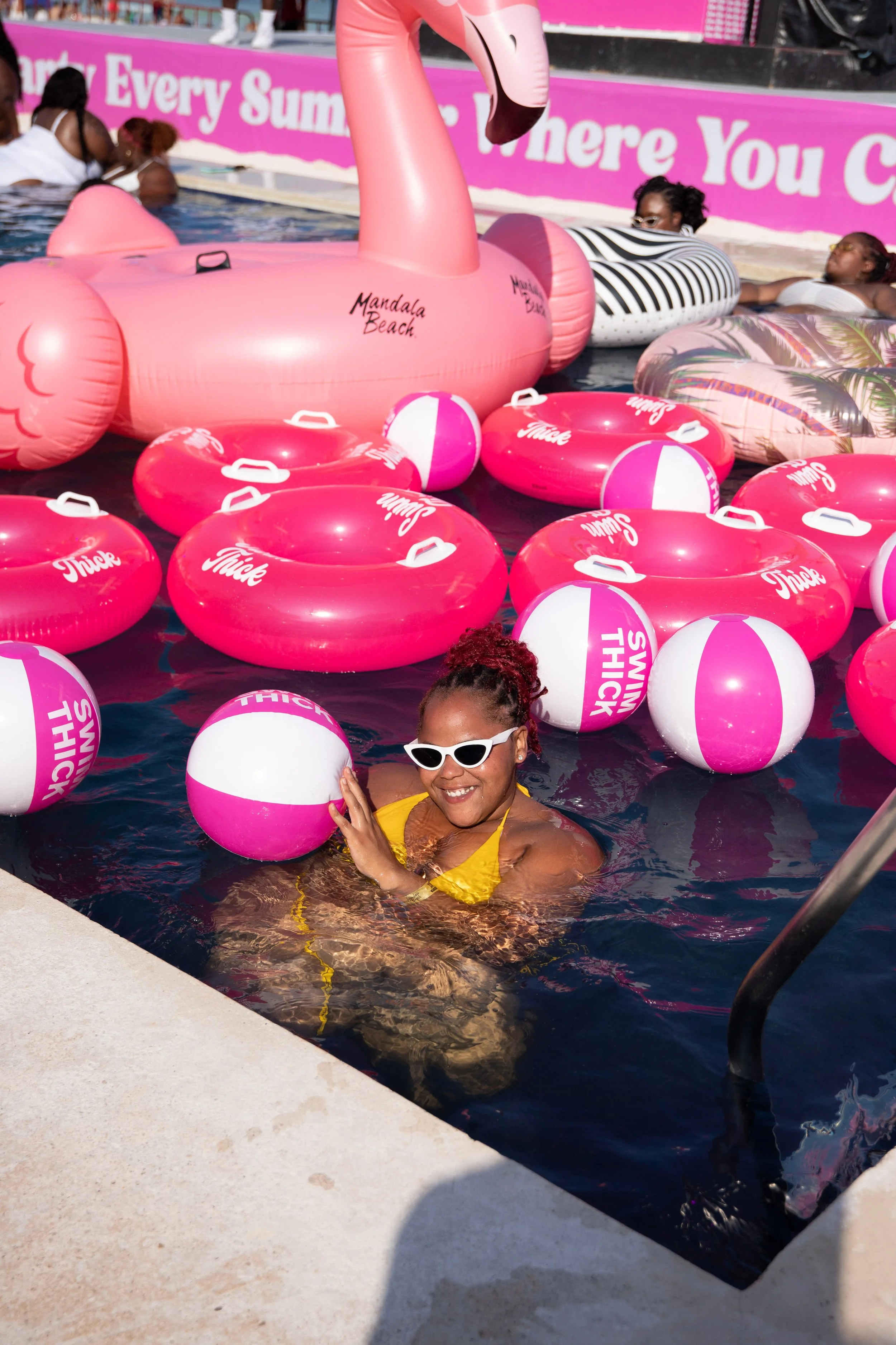 A woman smiling and wearing sunglasses, enjoying a swimming pool surrounded by pink and white beach balls and inflatable floats, including a pink flamingo, at Mandala Beach.