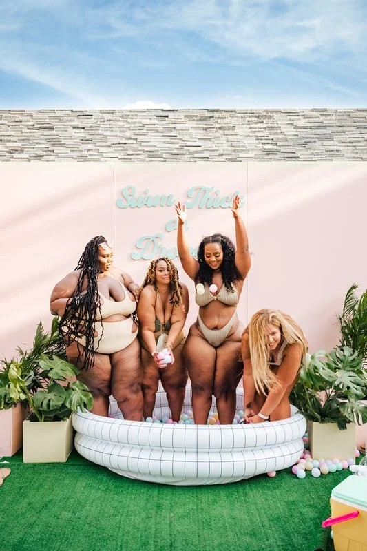 Four women in swimsuits playing with colored balls inside a small inflatable pool, behind a pink wall with a sign that says 'Sugar Thicc'.