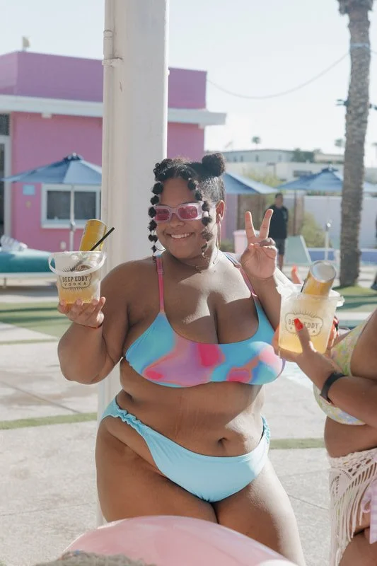 A woman in a colorful bikini smiling, holding a cocktail, and making a peace sign at an outdoor poolside area with pink and purple buildings in the background.