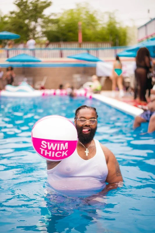Man with glasses and a beard in a pool holding a pink and white beach ball that says "Swim Thick," with other people and blue umbrellas in the background.