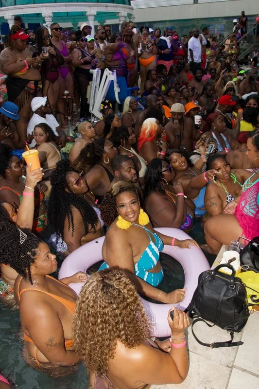 Large group of women enjoying pool party, some sitting in inflatable pools, others standing by poolside with drinks, wearing swimsuits and accessories, at an outdoor venue with a building in the background.