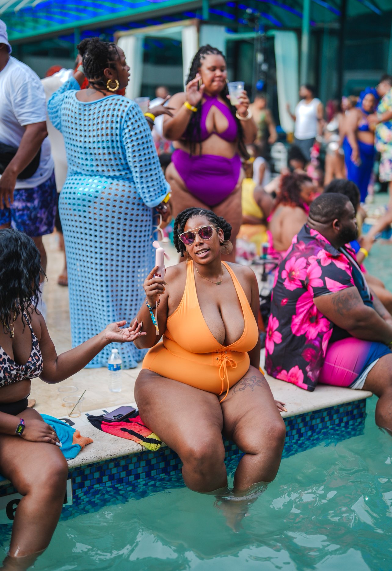 People enjoying a pool party outdoors, with women in swimsuits, some sitting on the pool edge with their feet in the water, others standing and mingling, colorful clothing and drinks visible.