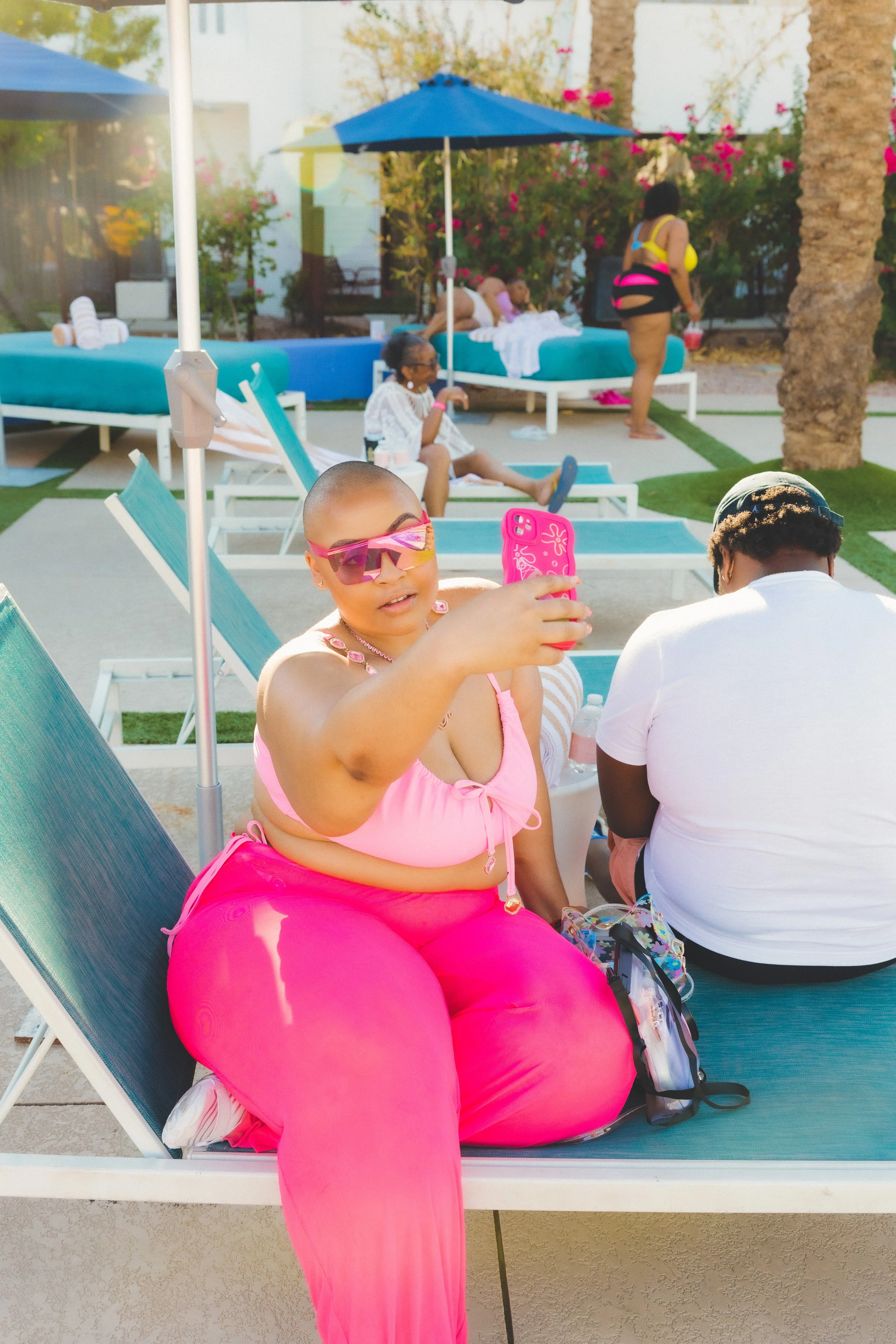 A woman in pink clothing taking a selfie by a poolside lounge chair, with other poolside visitors relaxing in the background.