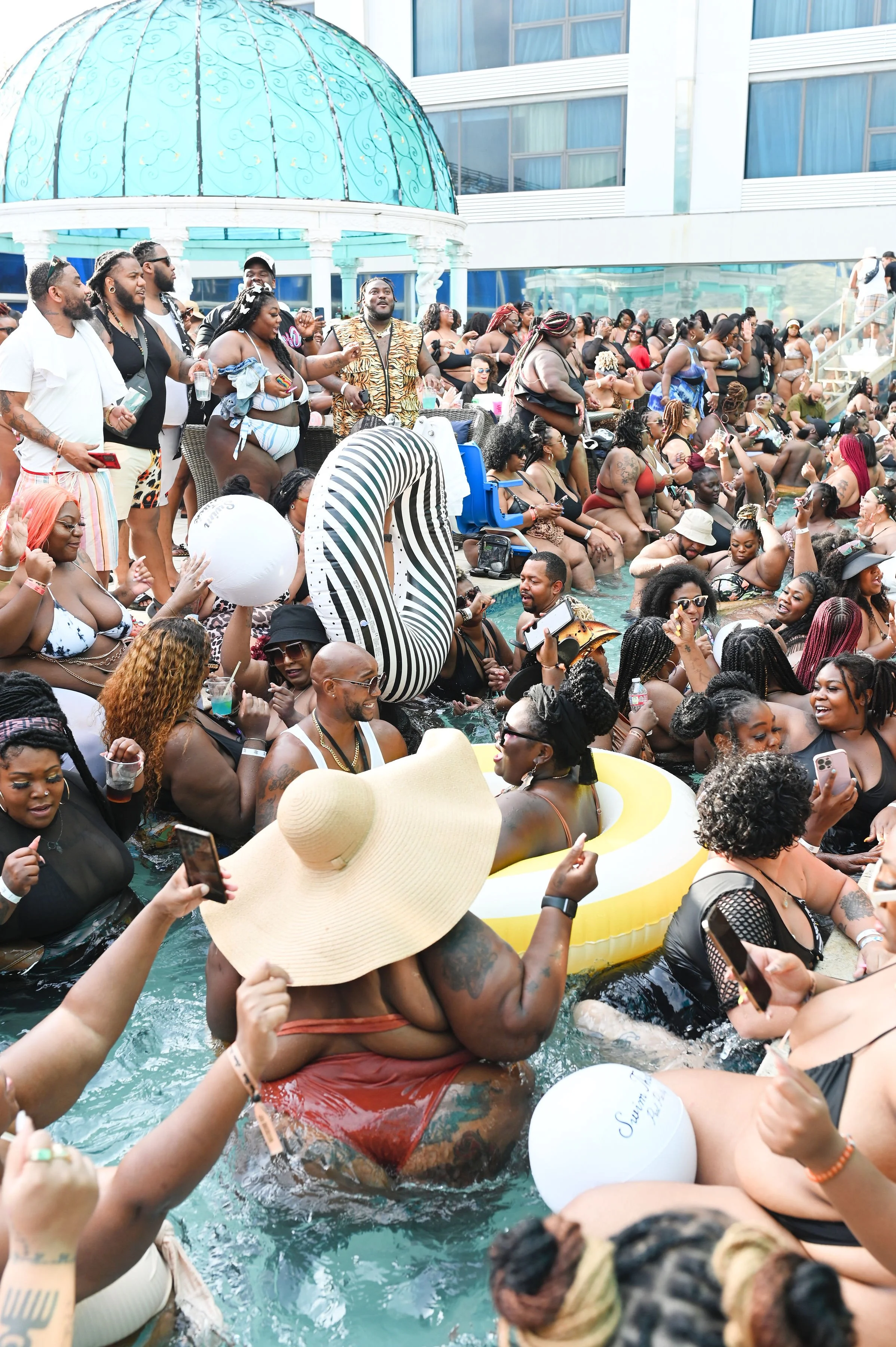 Crowd of people enjoying a pool party on a rooftop, with some wearing swimsuits and accessories like hats and sunglasses, some in the pool and others gathered on the pool deck.