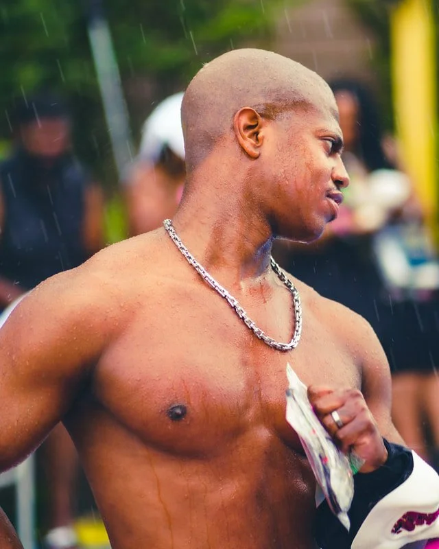 A shirtless man with a shaved head wearing a silver chain necklace, holding a cloth, during a rainy outdoor event.