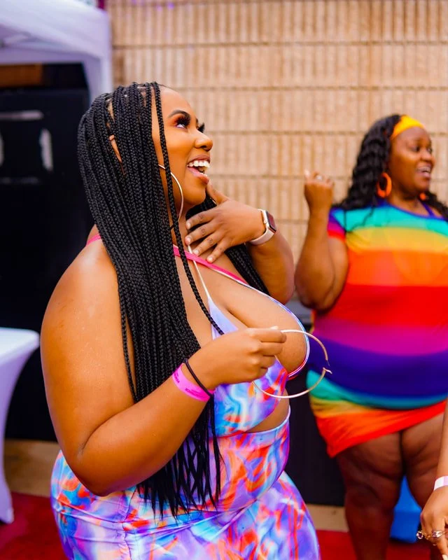 Two women at a joyful gathering, one with long braided hair wearing a colorful dress with a rainbow pattern, smiling and holding jewelry, the other woman in the background wearing a rainbow-striped dress, also smiling and celebrating.
