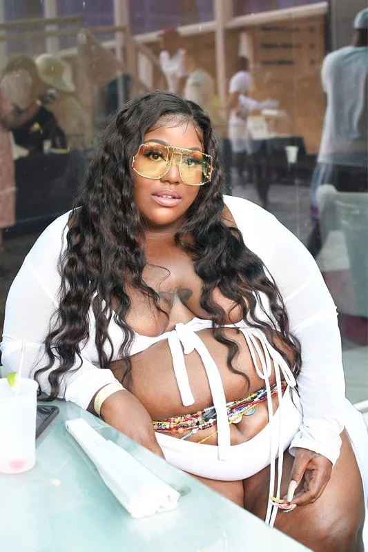 A woman with long curly hair wearing large yellow sunglasses, a white tied crop top, colorful beaded accessories, and a white jacket, sitting at a table in a casual indoor setting.