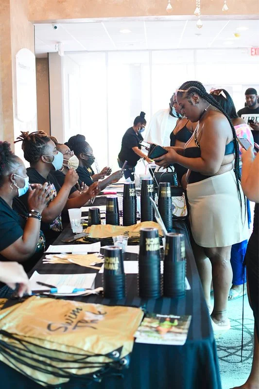 Group of women wearing face masks sitting at a table with filled with black cups, brochures, and bags, interacting with a standing woman in a black top and white skirt in a brightly lit indoor space.