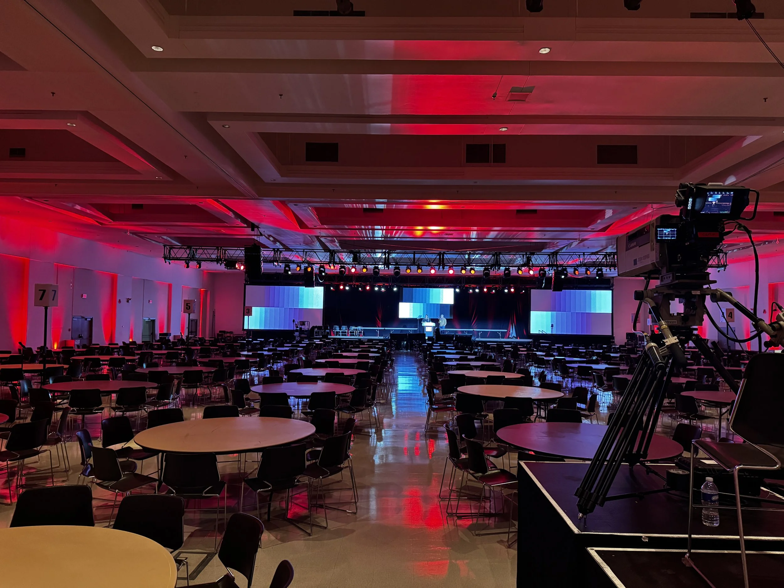 Empty conference hall with round tables, black chairs, a stage with large video screens, colorful red and blue lighting, and cameras set up for recording or broadcasting.