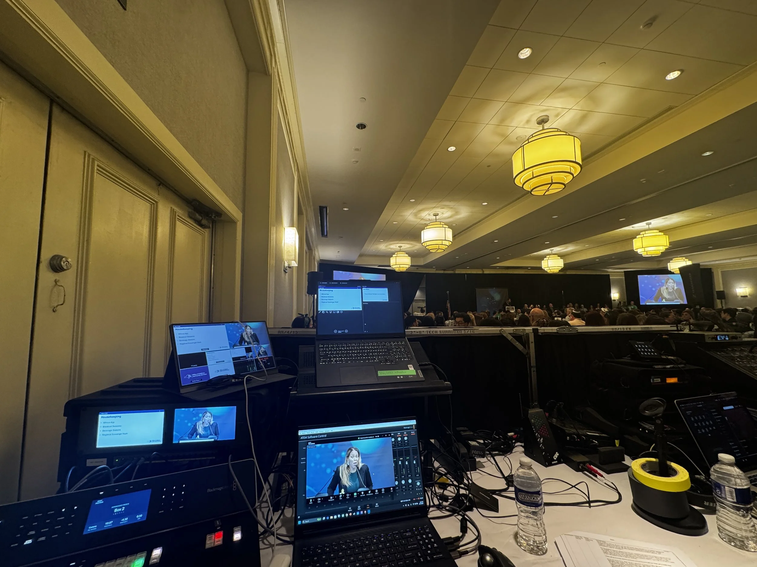 View of a conference room with a stage and large screen at the front, filled with attendees, as seen from the technical control area with multiple monitors and equipment in the foreground.
