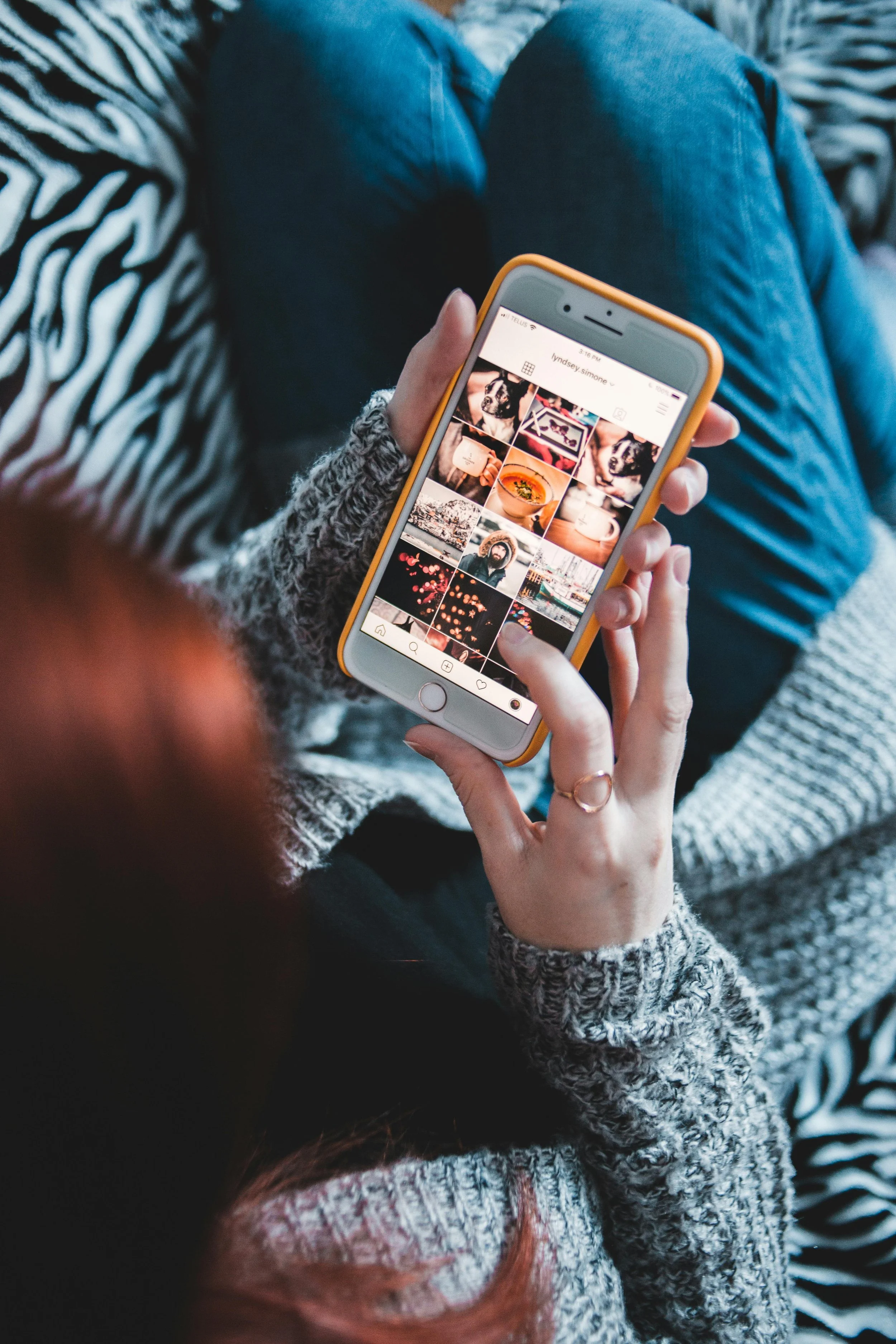 Person in a gray sweater holding a smartphone browsing Instagram feed with images in a cozy setting.