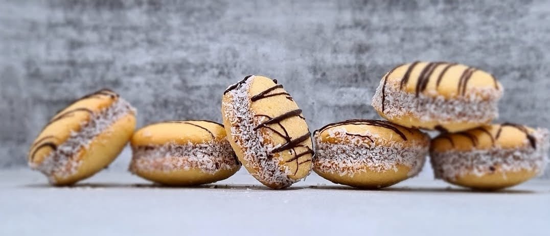 A row of six alfajores with chocolate drizzle, and shredded coconut, set against a blue textured background.