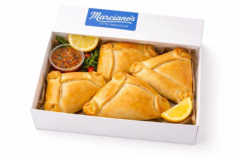 Four golden brown baked empanadas in a white box with a blue and white label saying marcianos  , garnished with lemon wedges, fresh herbs, and a small container of pebre dipping sauce.