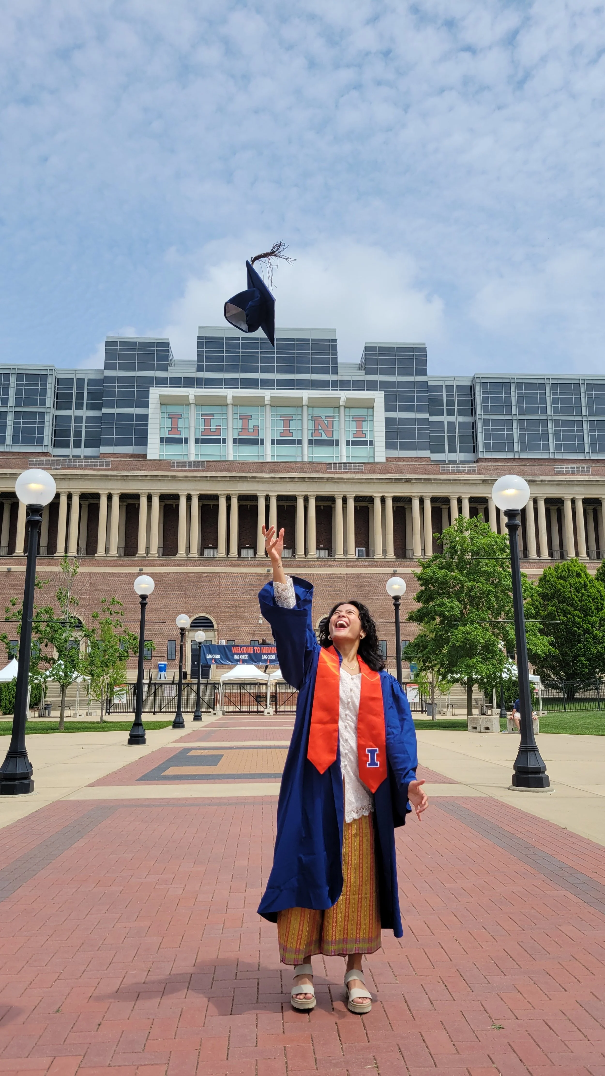 Audrey is wearing a traditional Indonesian get up with a white blouse and colorful sarong, white heels, a dark blue graduation robe, and orange banner with a blue "I." She is tossing her blue graduation cap in the air. The stadium is behind her.