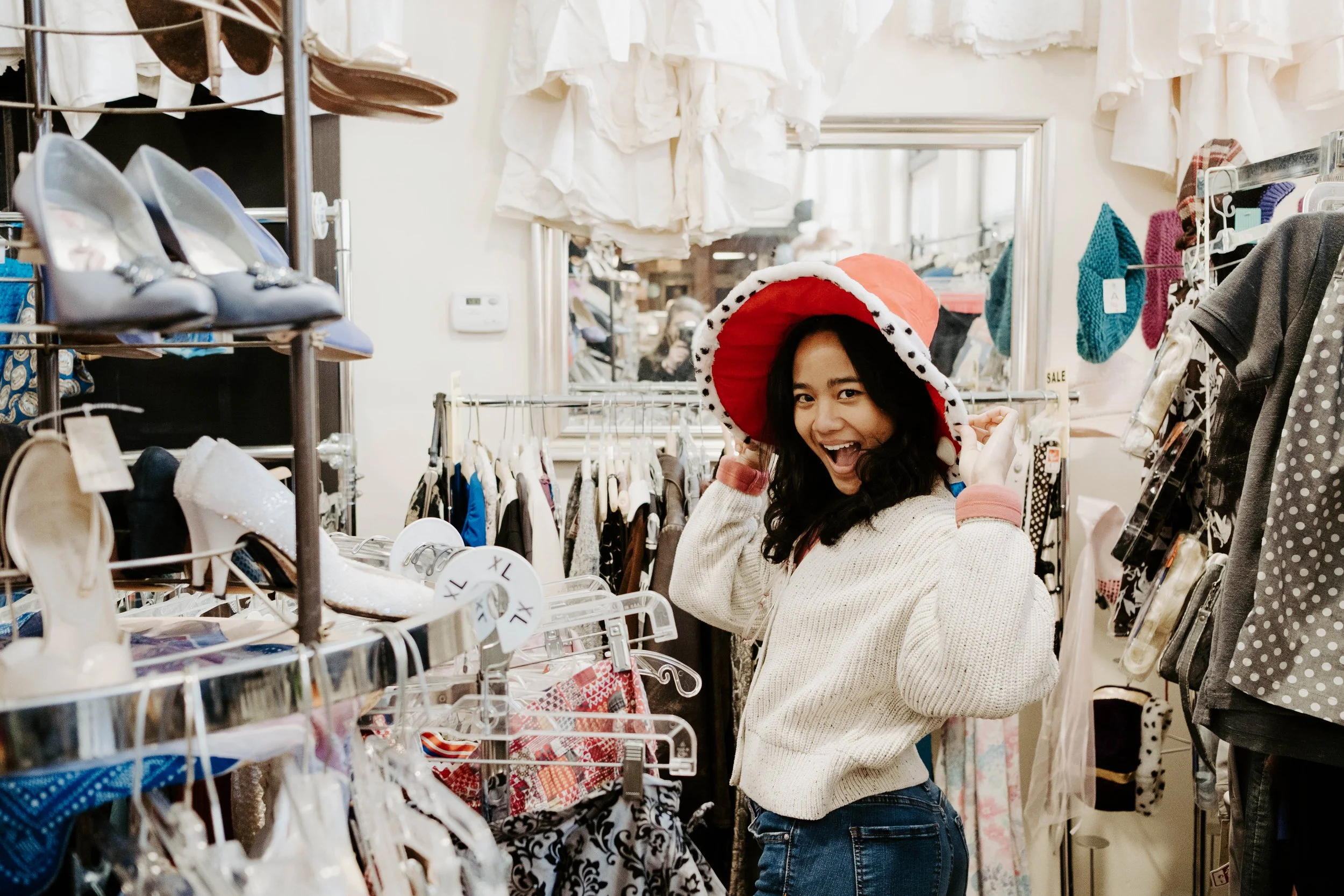 Audrey is wearing white cardigan, jeans, and red hat with white brim with black polka-dots. She is smiling with her hands on the hat to the camera. She is in a thrift store with tons of clothes on racks in the background.