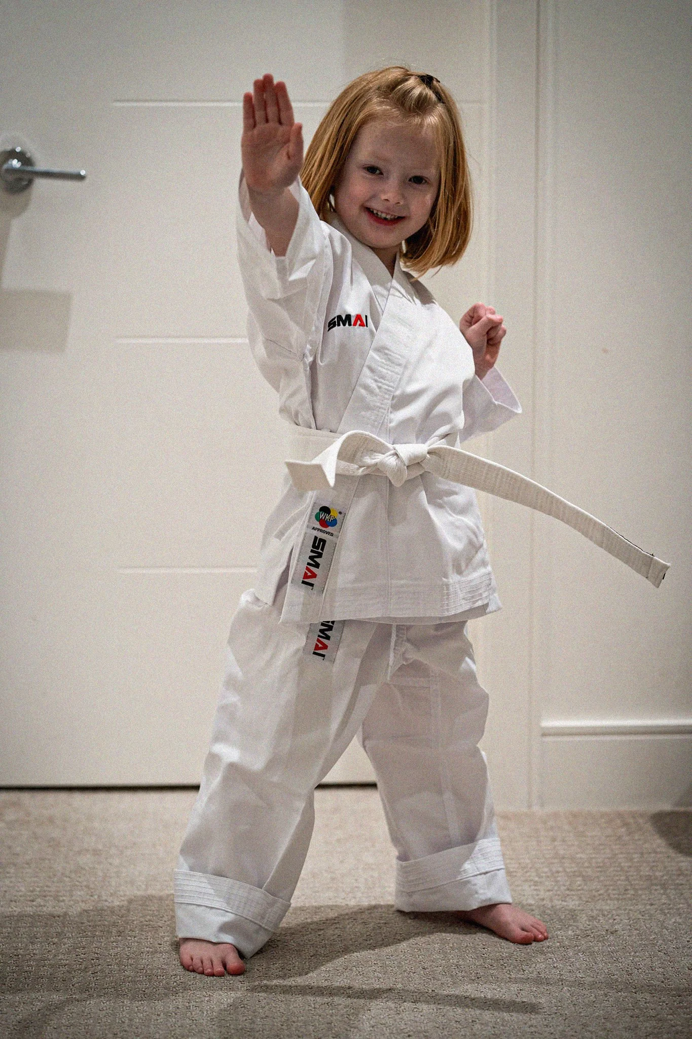 A young girl with red hair wearing a white martial arts uniform and a white belt, smiling with her fist up.