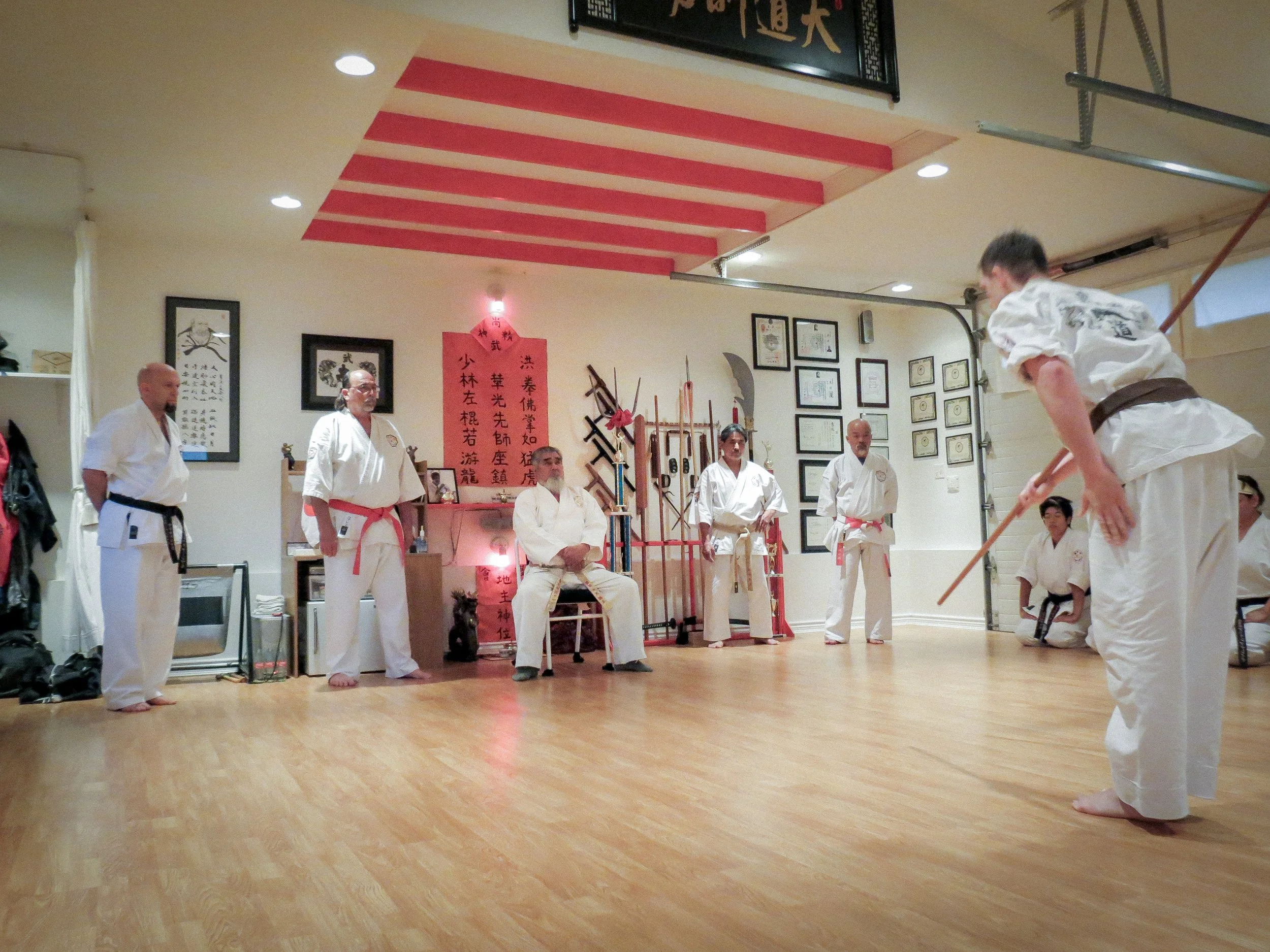 Martial arts demonstration in a dojo with students and instructor, some holding weapons, one student bowing holding a staff.