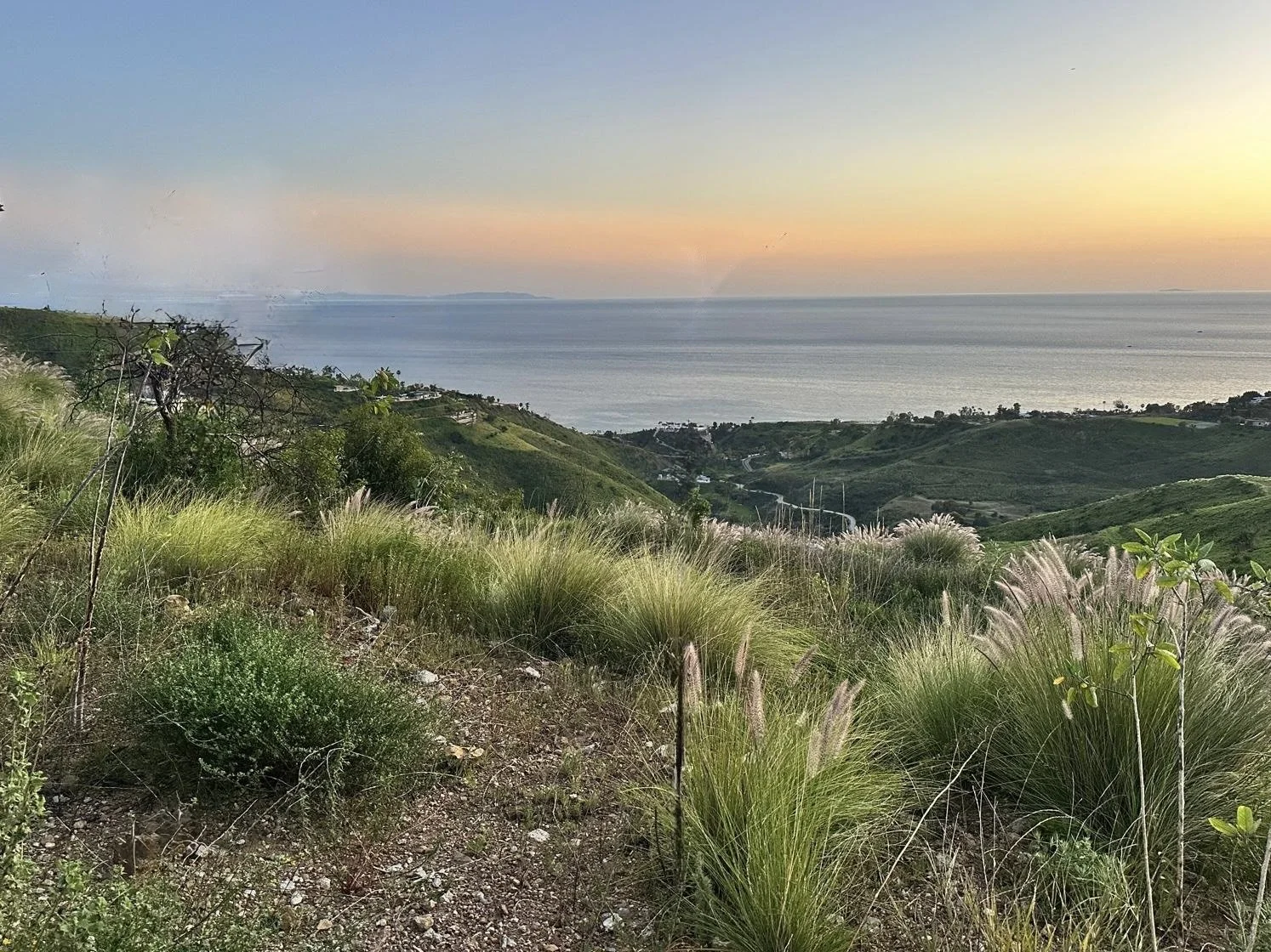 Sunset over hills and ocean, with grassy foreground and distant land on the horizon.
