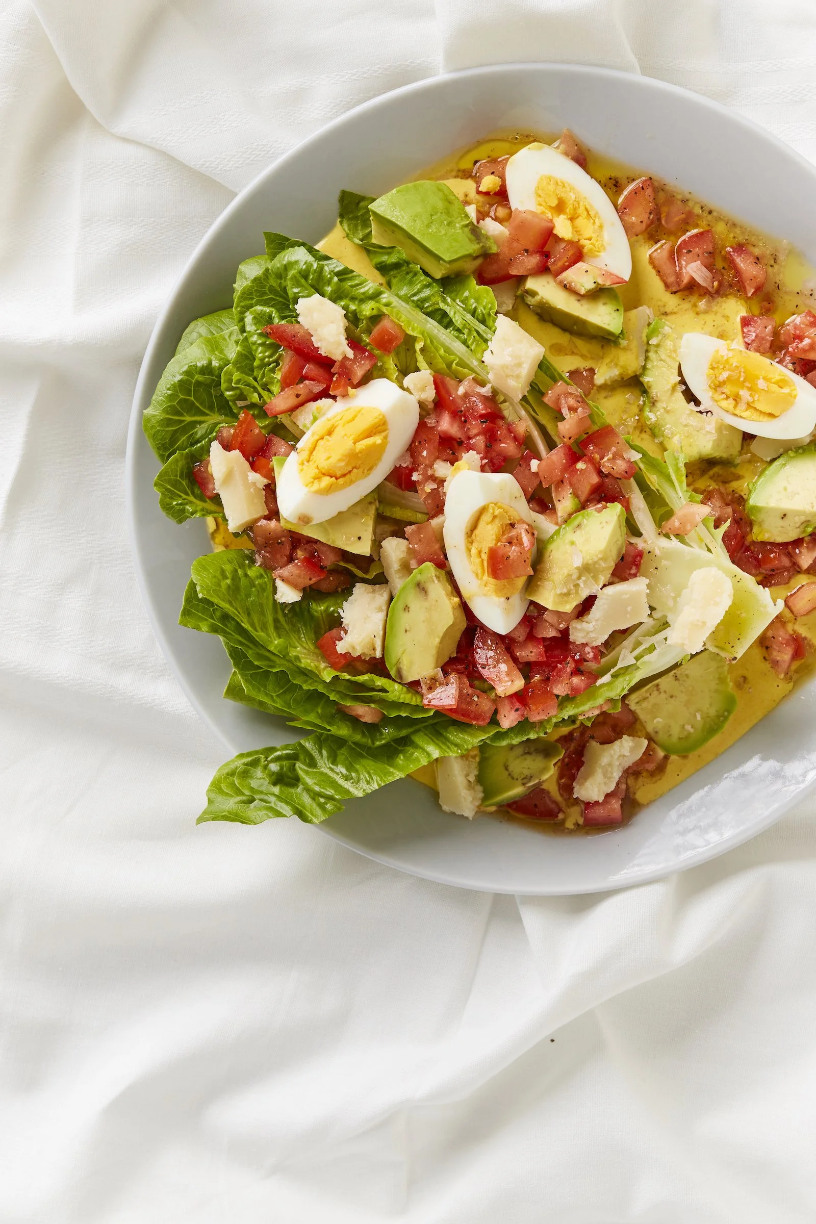 A bowl of salad with lettuce, hard-boiled eggs, avocado, tomato, cheese, and ham, set on a white cloth.