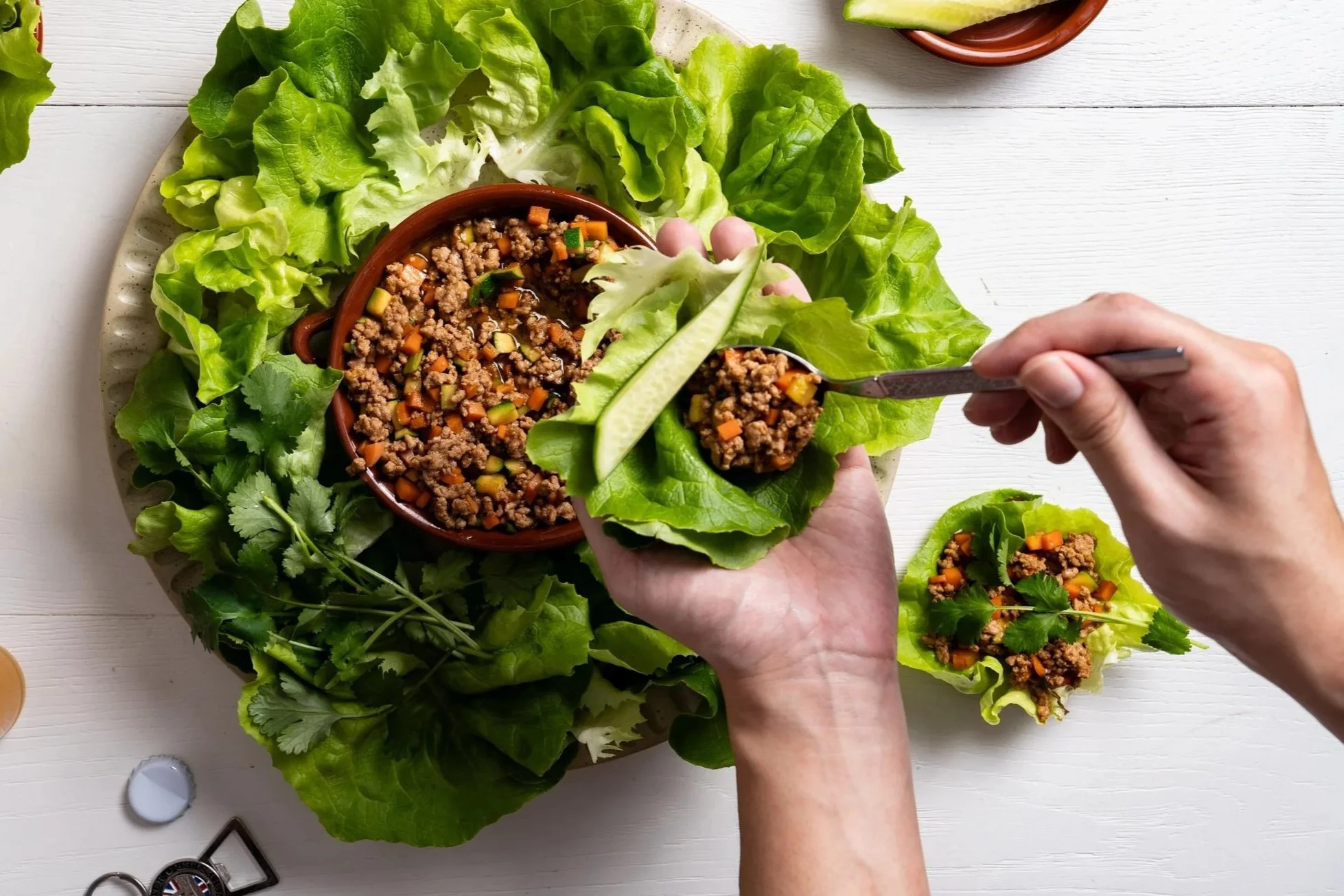 Person filling lettuce leaves with cooked ground meat, diced vegetables, and herbs on a white plate.