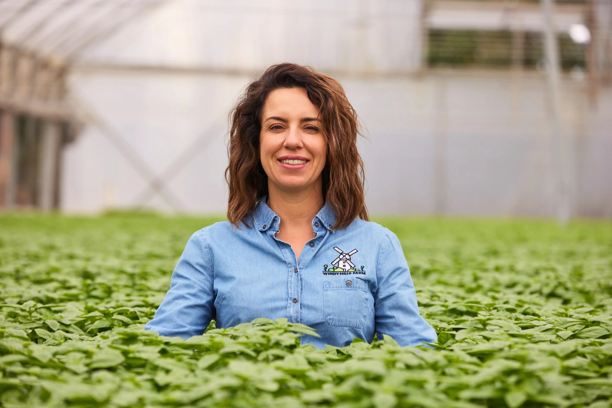 A woman with curly brown hair smiling in a greenhouse filled with green plants, wearing a blue shirt with a farm logo.