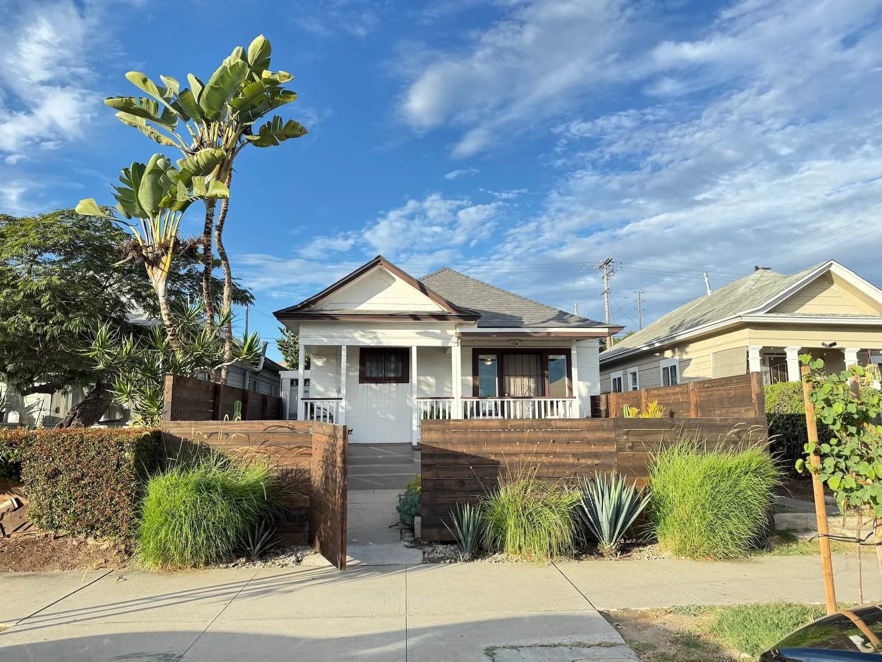 Front view of a small white house with a porch and a black door, surrounded by a landscaped yard with green plants, and a tall banana tree on the left, under a partly cloudy blue sky.