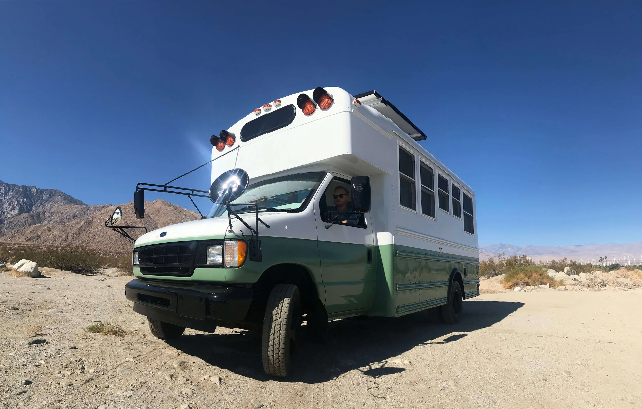 A converted school bus, also known as a Skoolie, parked in a desert landscape with mountains in the background and clear blue sky.