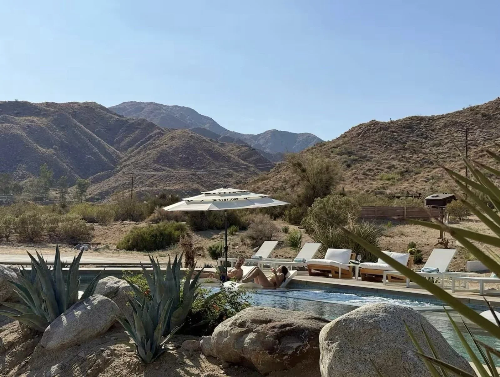 People relaxing in a luxury wellness property's pool with lounge chairs and umbrellas, surrounded by desert landscape and mountains.