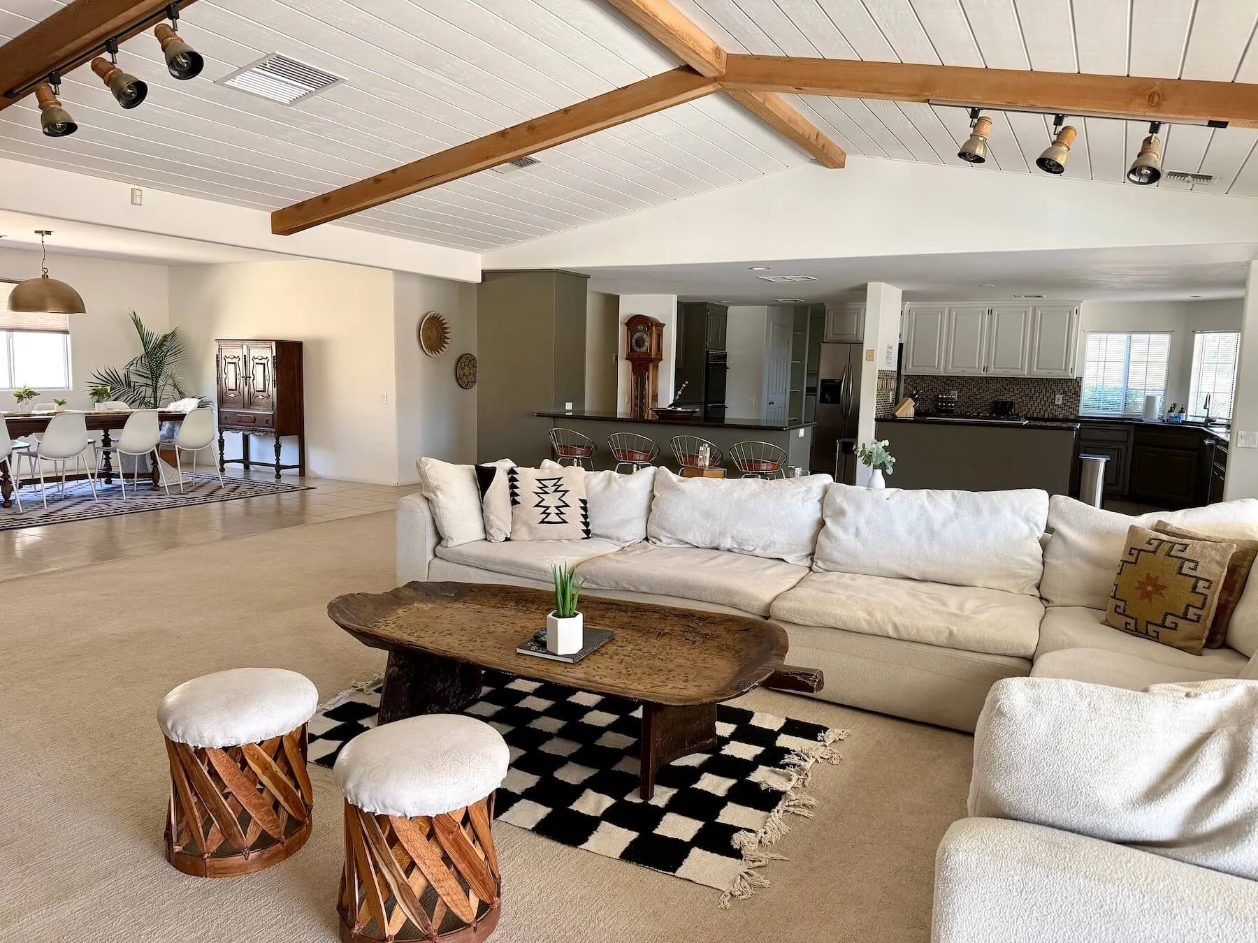 Living room with cream sectional sofa, black and white checkered rug, wooden coffee table, and two wooden stools with white cushion in front. Open floor plan featuring dining area with white chairs, and kitchen with white and dark cabinetry.