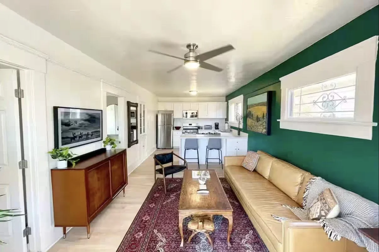 Living room with teal accent wall, beige sofa, wooden coffee table, patterned rug, and kitchen with white cabinets and stainless steel appliances.