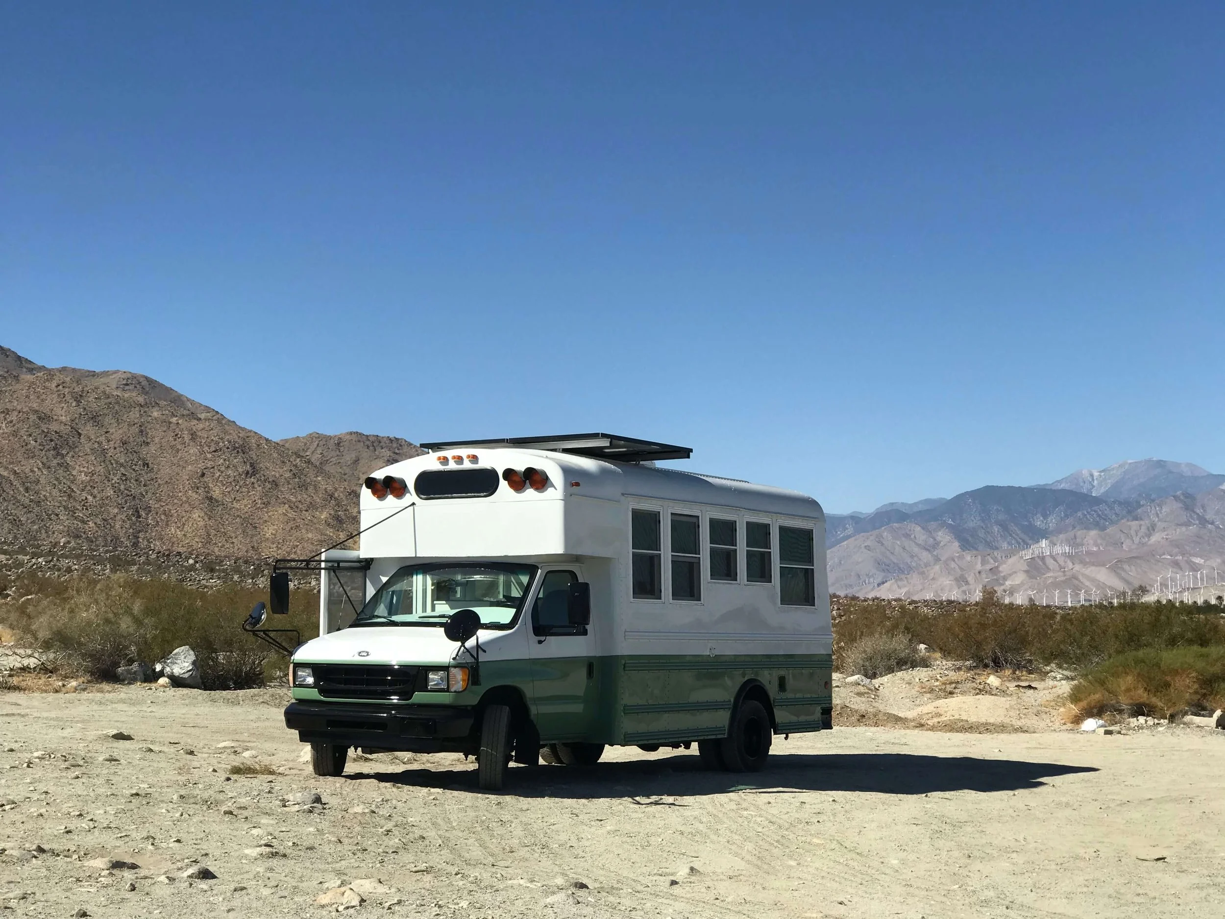 A white and green Skoolie parked on a dirt area in a desert landscape with mountains in the background and wind turbines on the horizon.