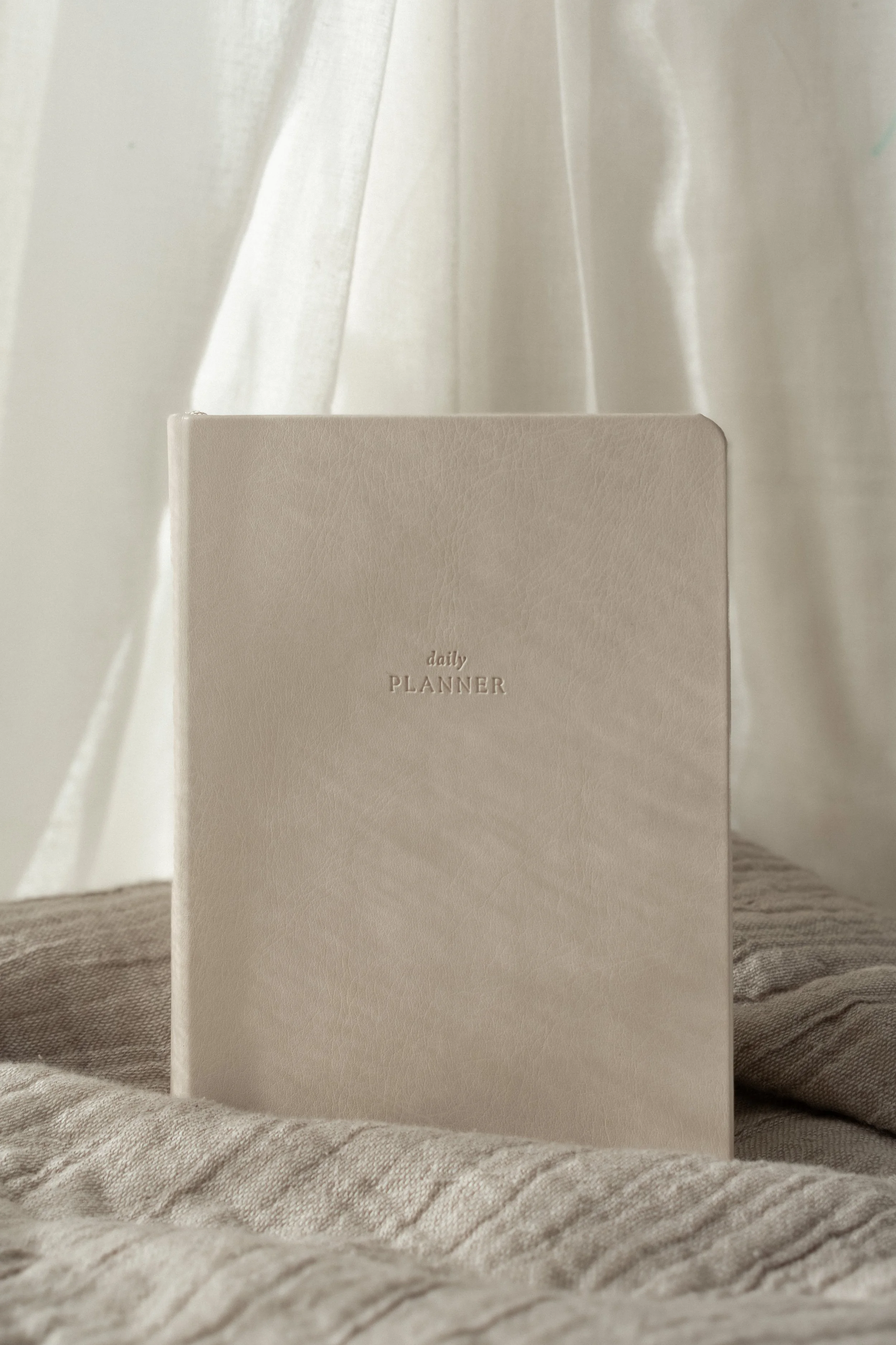 An off-white vegan leather-bound daily planner standing upright, with a light-colored curtain in the background.