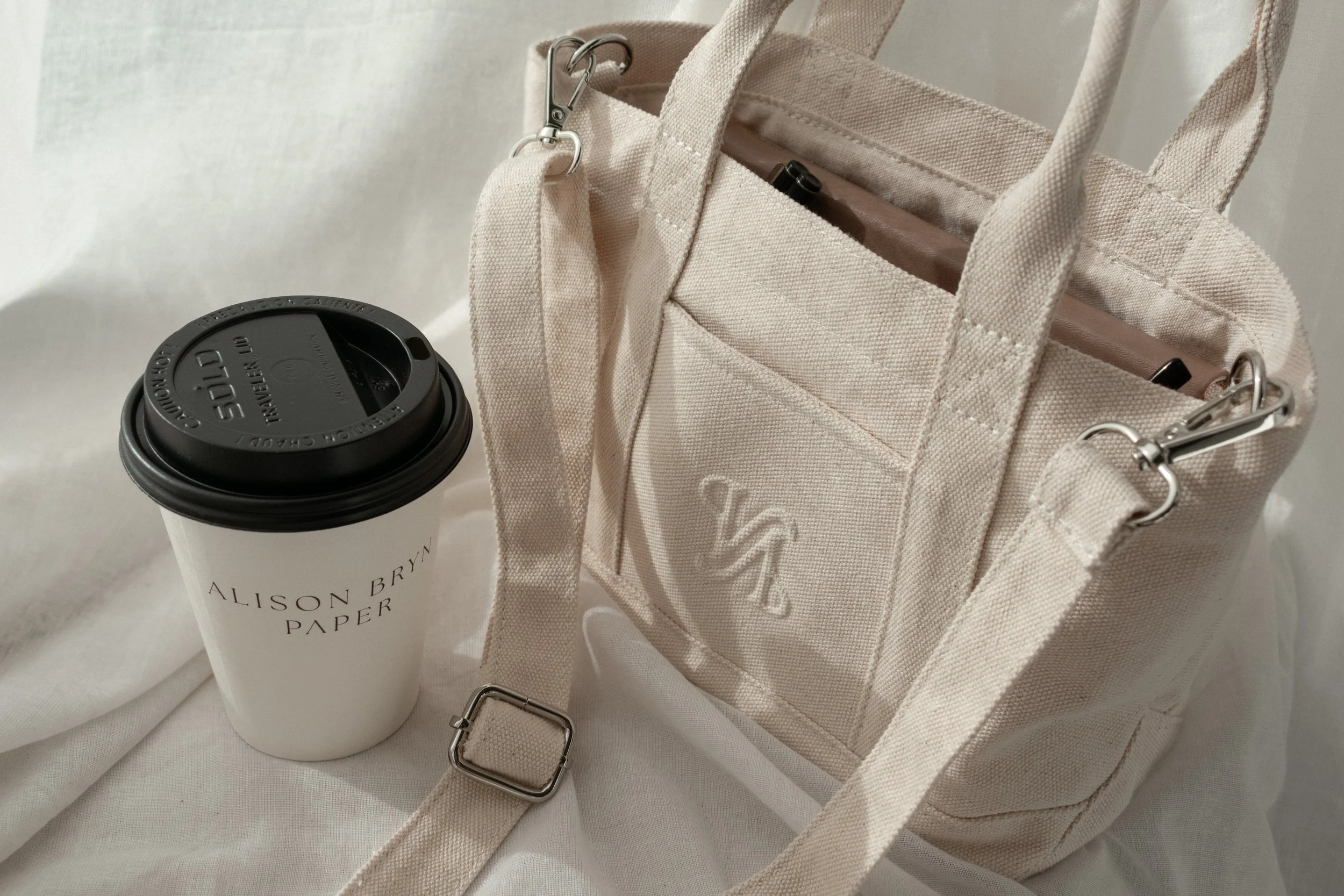 Beige canvas bag with a strap and a logo on the pocket, a white paper coffee cup with a black lid and printed text, and a white fabric background.