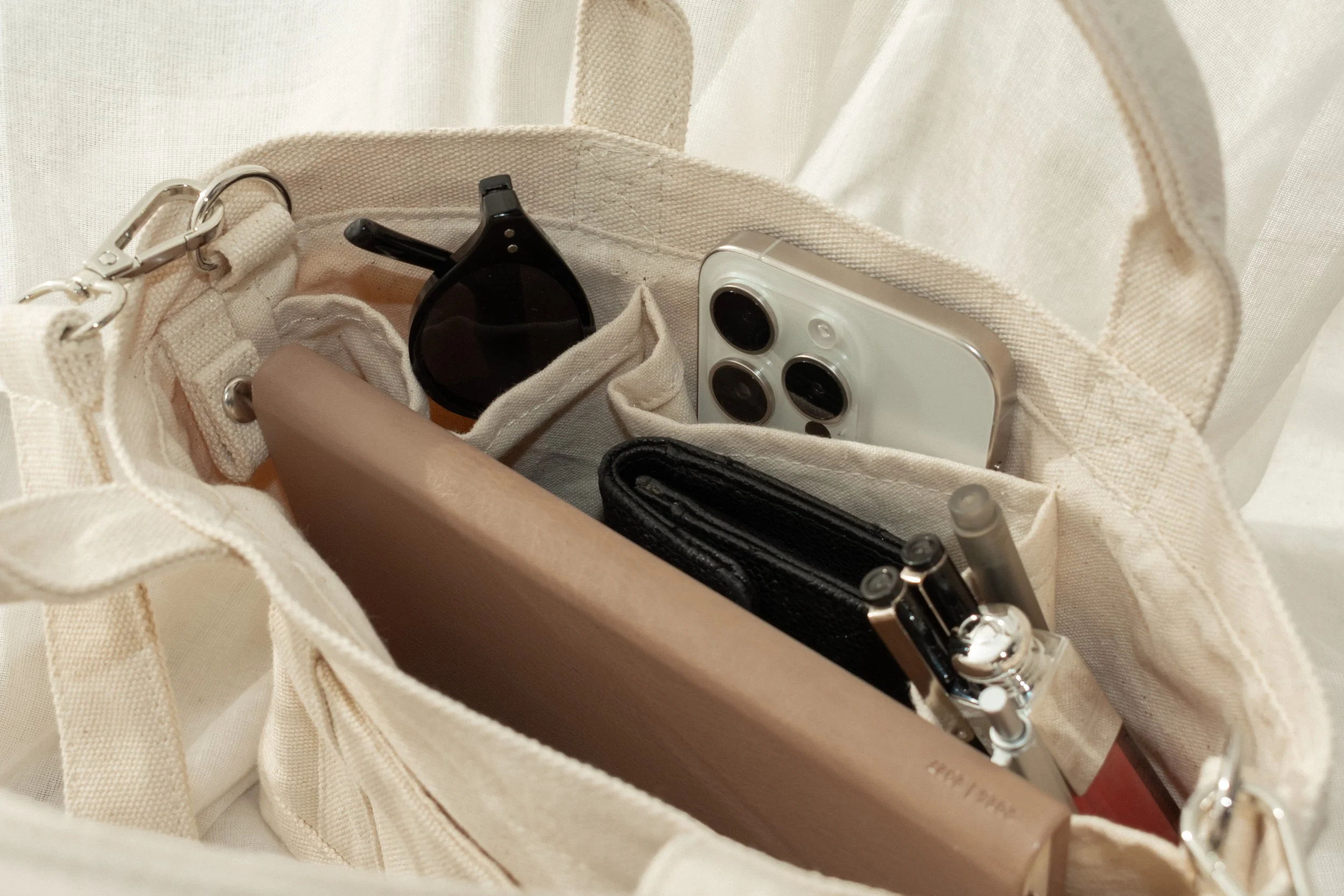 Open beige tote bag containing sunglasses, a white smartphone, a black wallet, pens, and a beige notebook.