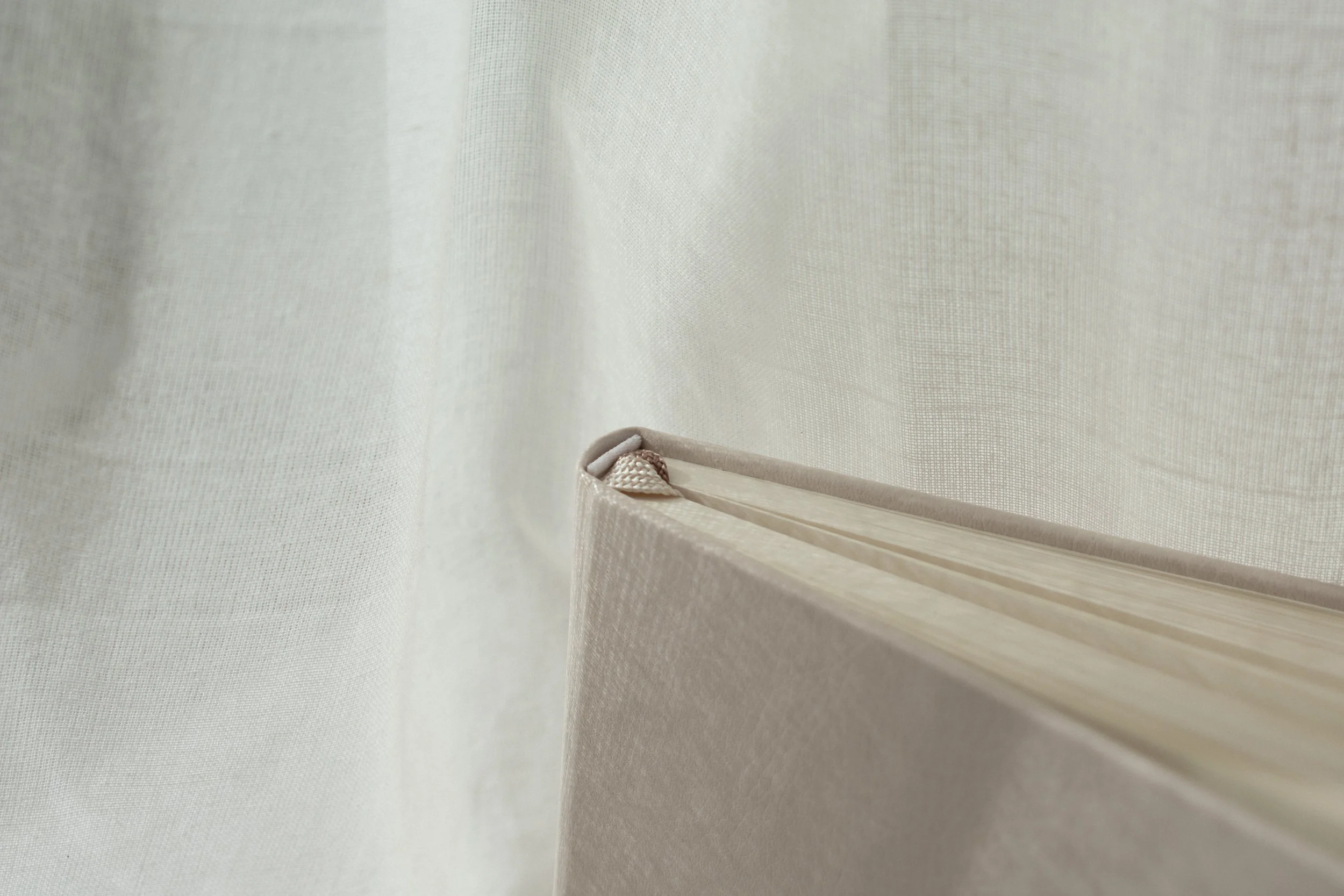 Close-up of a beige fabric curtain partly pulled back to reveal a beige hardcover vegan leather planner with a fabric bookmark inside.