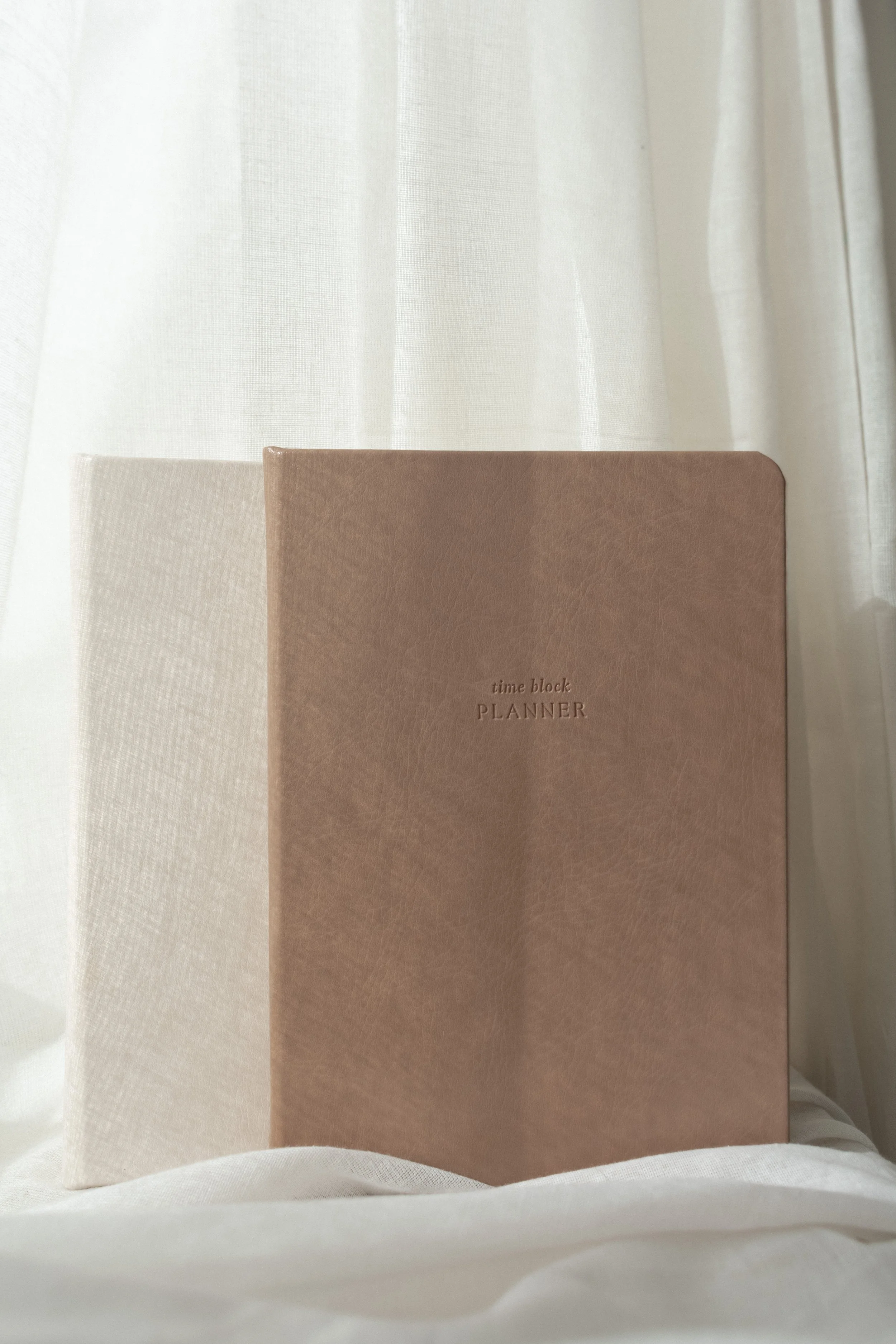 A brown vegan leather planner with 'time block PLANNER' embossed on the cover, sitting in front of a white fabric backdrop and partially covered by a sheer white curtain.
