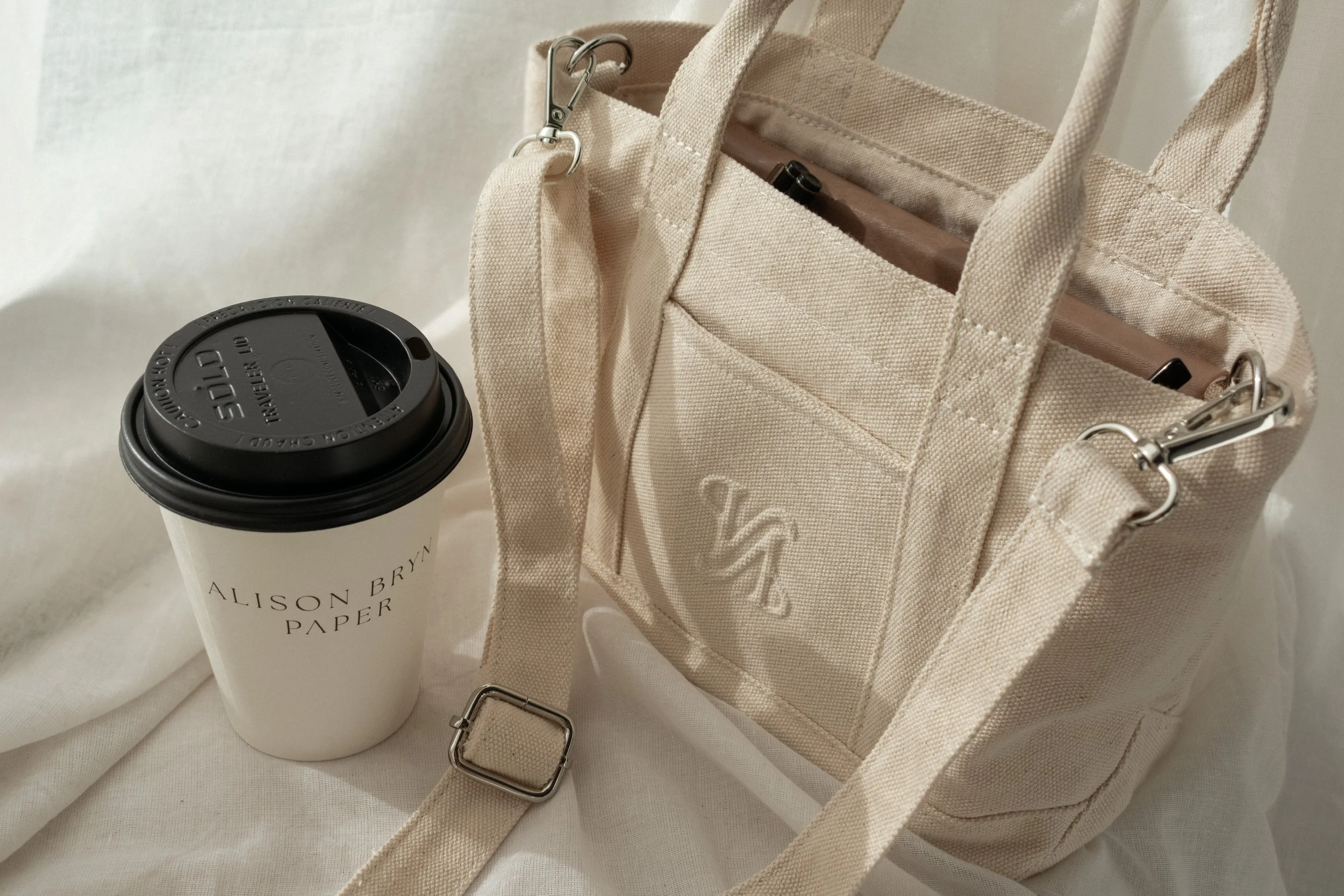 Beige canvas bag with a strap and a logo on the pocket, a white paper coffee cup with a black lid and printed text, and a white fabric background.