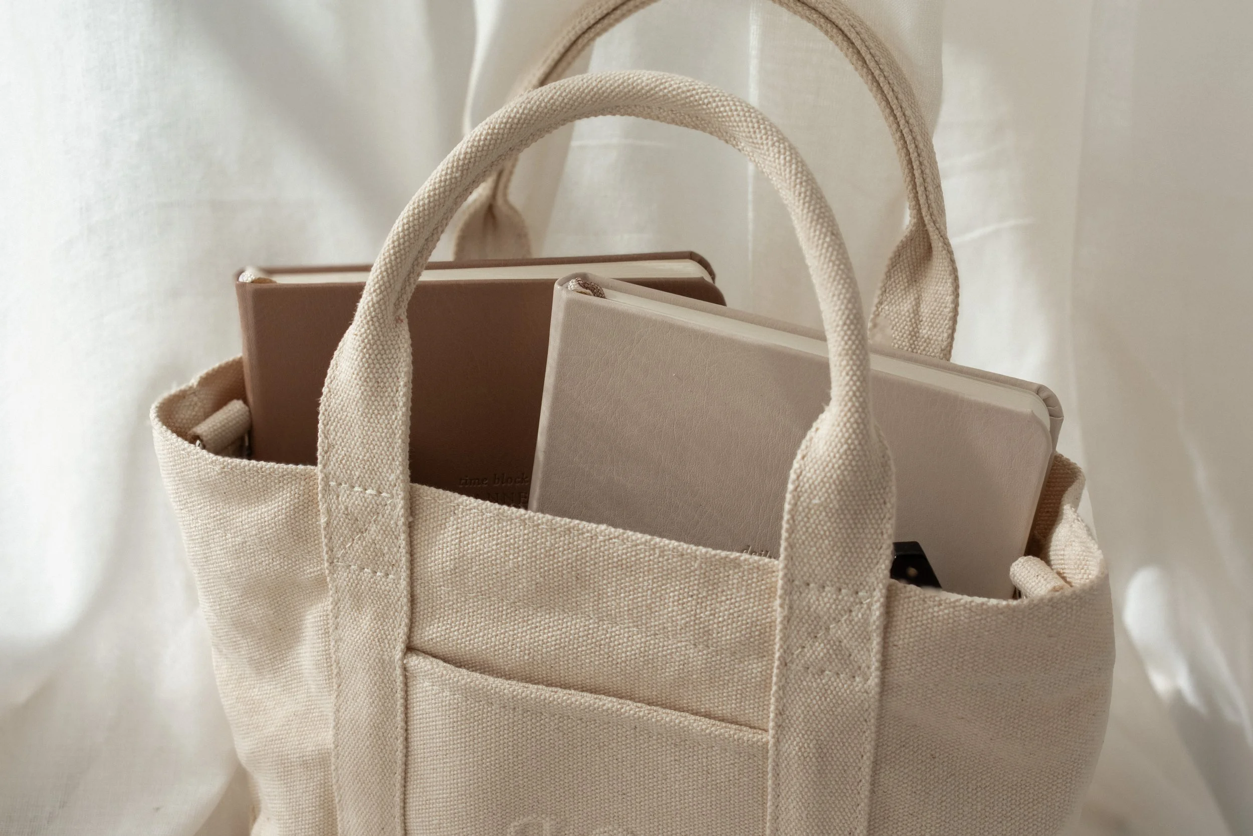 A beige canvas tote bag containing two books, one brown and one cream, set against a neutral fabric backdrop.