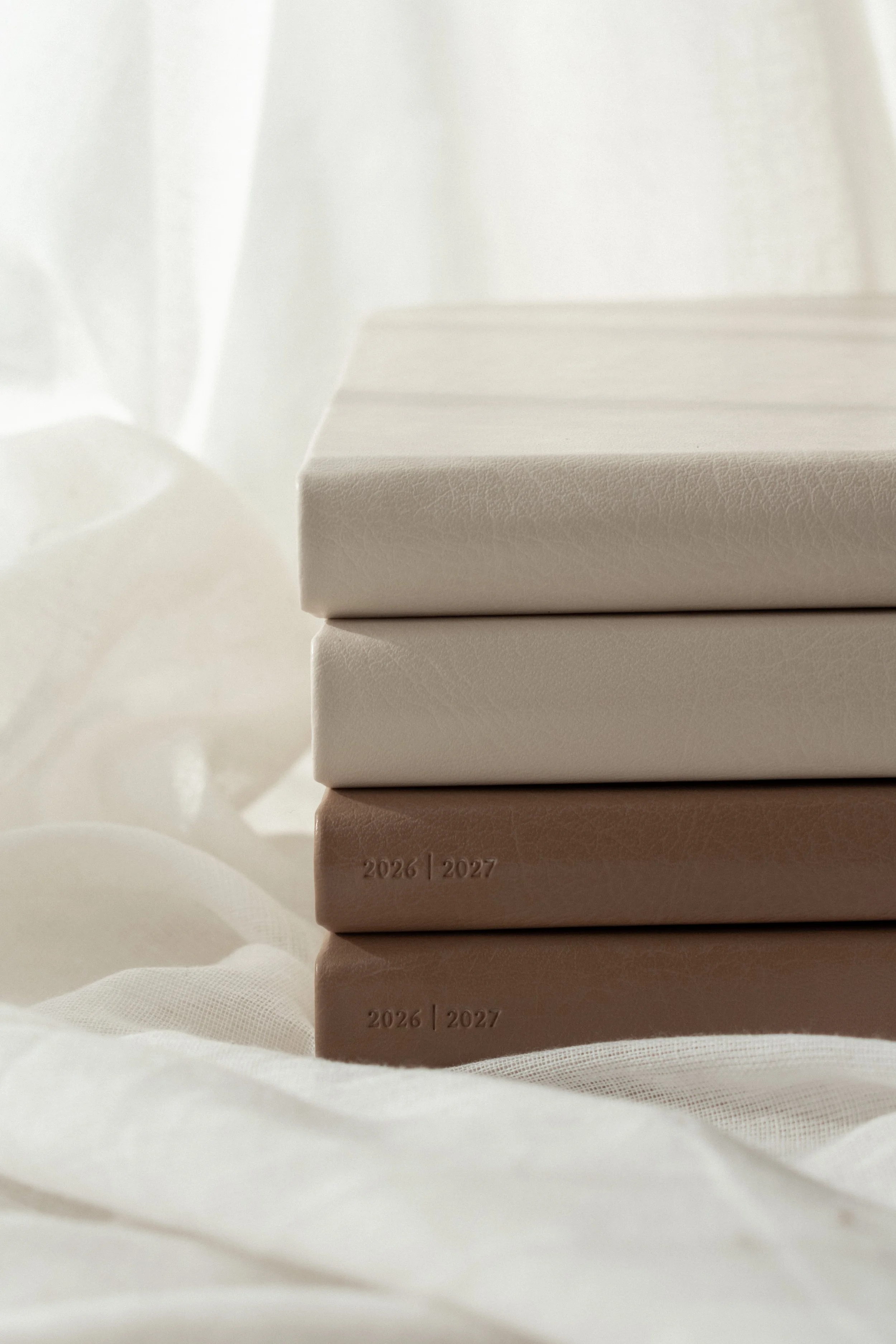 Stacked white and beige notebooks with embossed years 2026 and 2027, on soft white fabric with sheer curtains in the background.