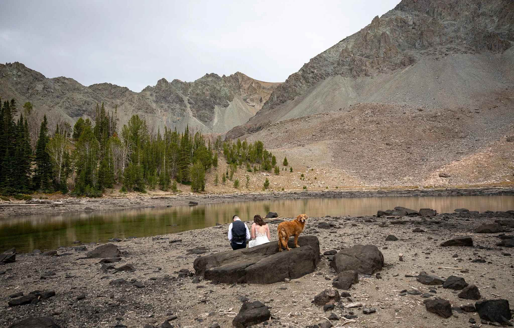 A couple with a dog sitting on a rocky shore by a lake, surrounded by mountains and pine trees under an overcast sky.