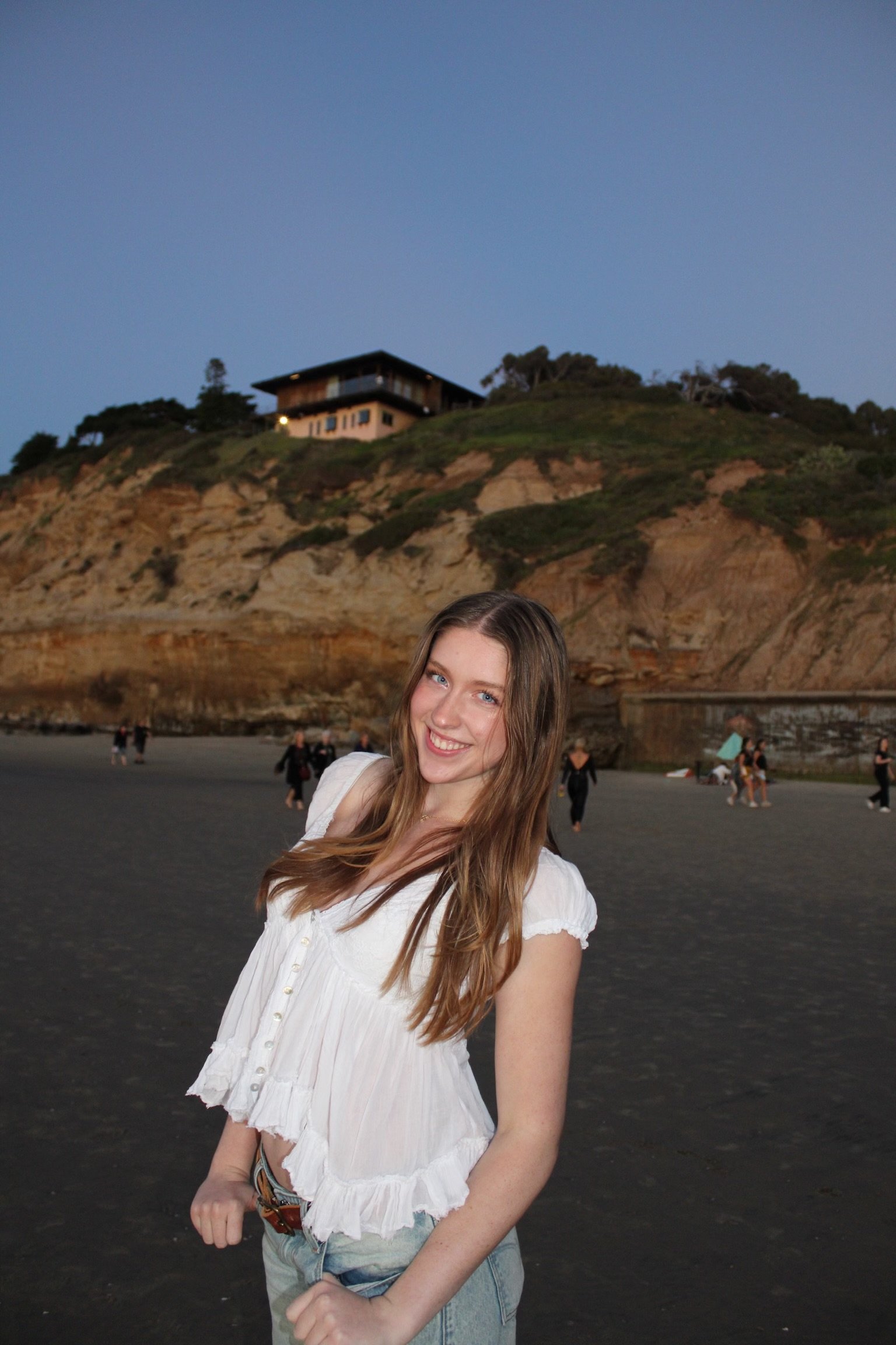 Image of a woman with long dirty blonde hair wearing a white blouse outdoors near the beach