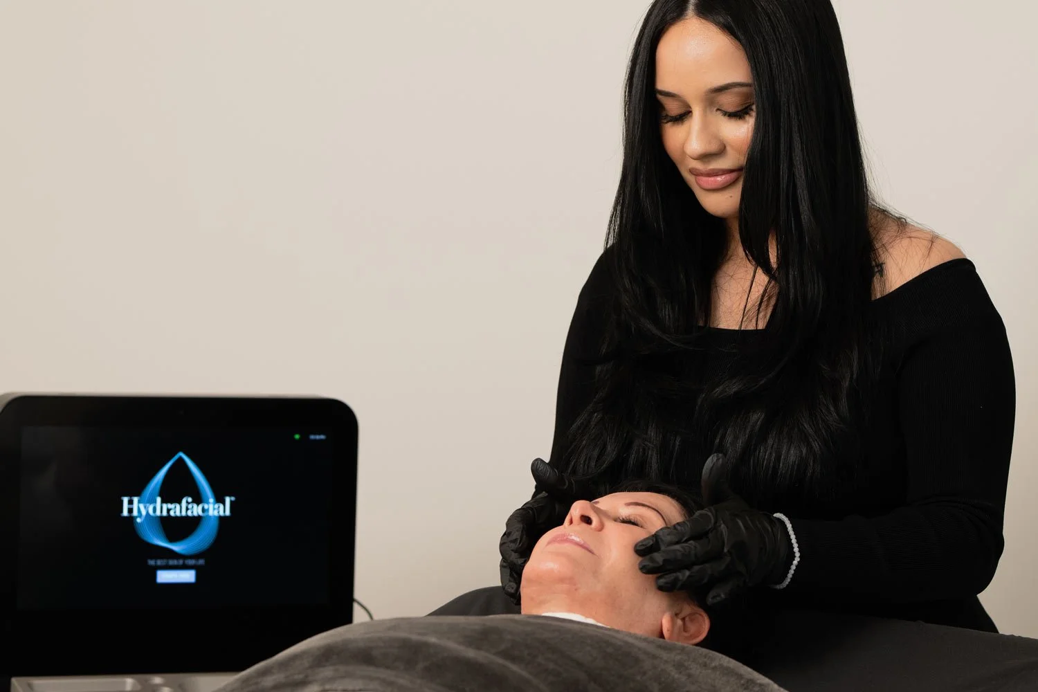 A woman is performing a HydraFacial on a client lying down with eyes closed, while a machine labeled 'Hydrafacial' is nearby.