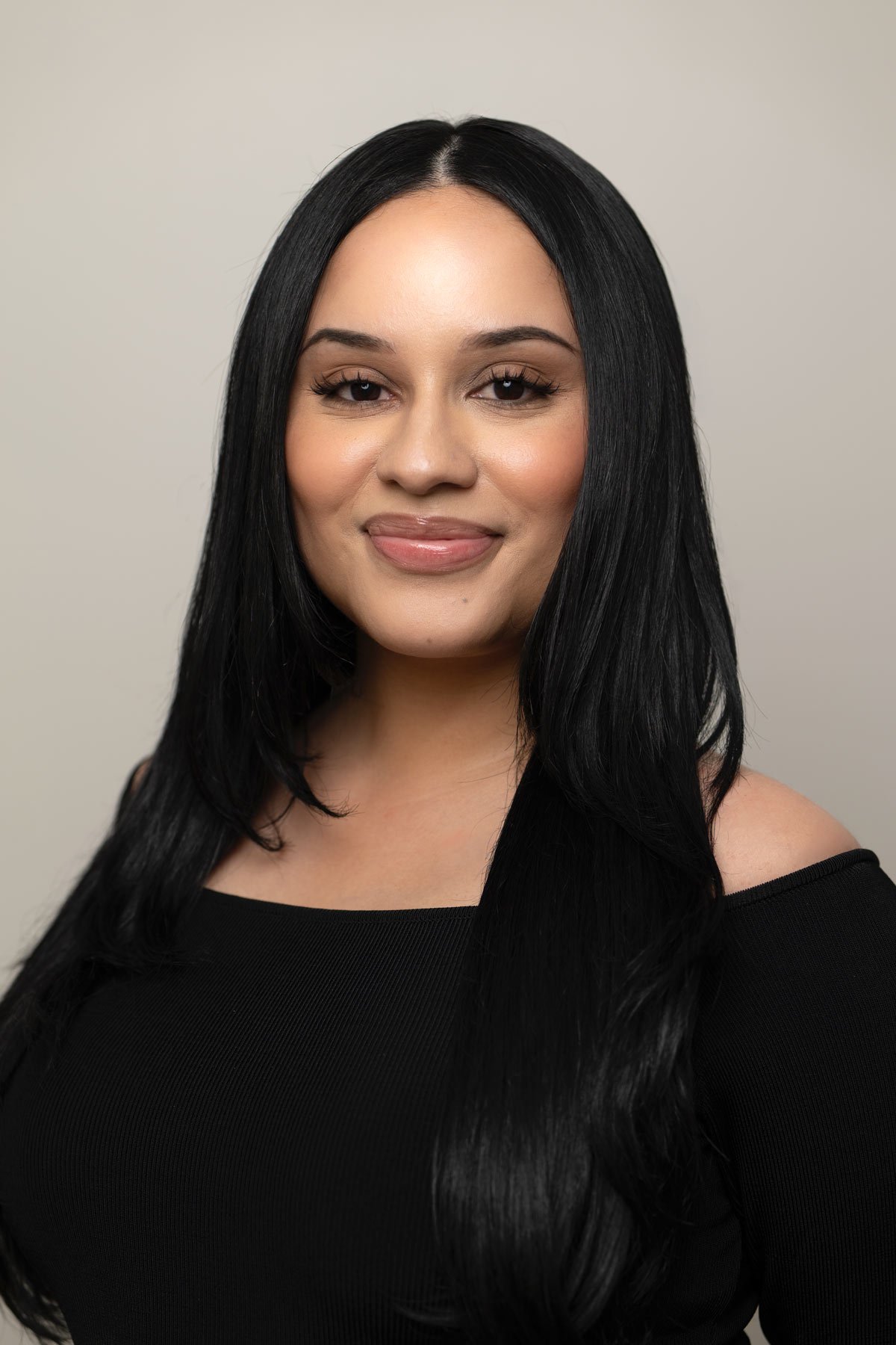 A woman with long black hair, wearing a black top, smiling at the camera against a plain beige background.